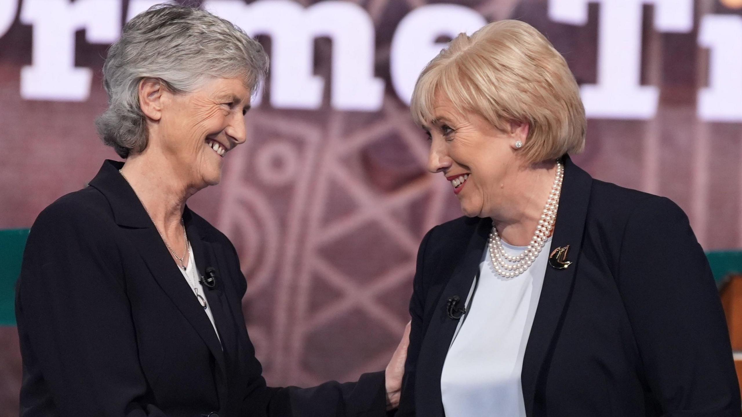 Catherine Connolly wearing a black suit and white top. She has grey hair. She is smiling at Heather Humphreys, also wearing a black suit and white top with a pearl necklace. She has blonde hair. They are smiling.