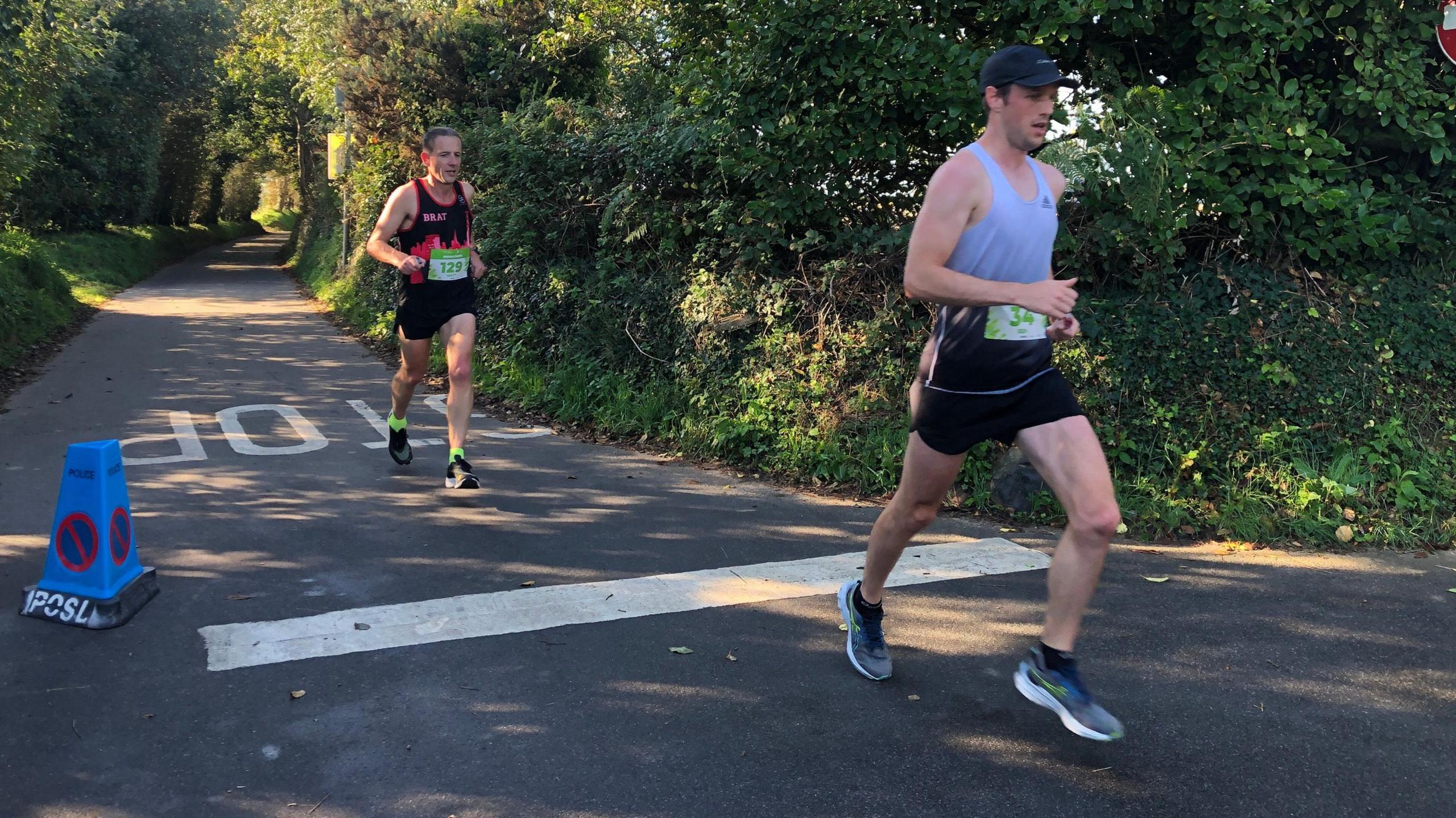 "Two runners on a sunlit paved road surrounded by dense greenery, participating in a race. The runner in front wears a light gray tank top, black shorts, and a cap, while the runner behind wears a black tank top and shorts. Both have race bibs. A blue traffic cone labeled 'IPSL' and a no parking sign are visible on the left. White road markings include an arrow and partial text '10'