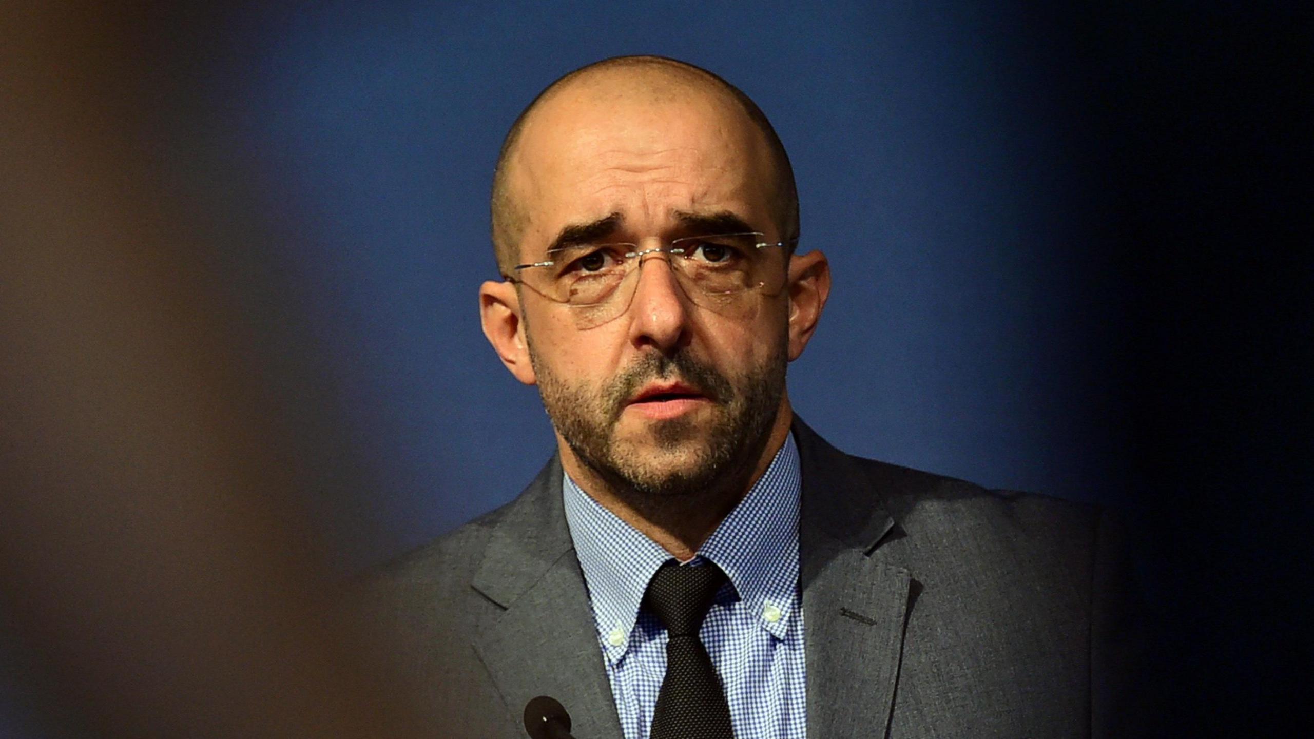 A man is looking towards the camera. He wears glasses and has a grey suit on. Something is partially obscuring the front of the photo. 