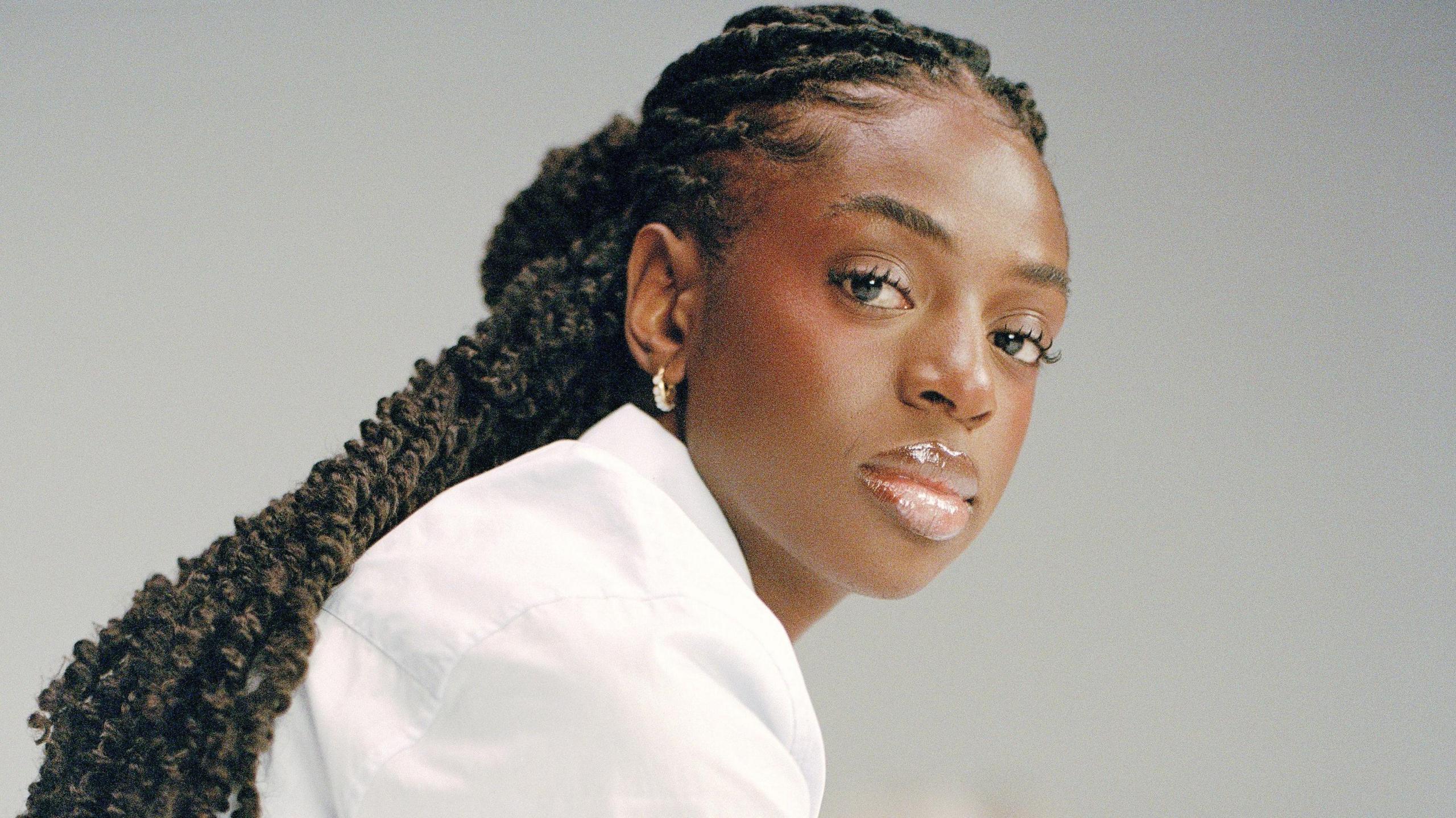 Cat Burns in a promotional shoot. She wears a white shirt and has her braided hair tied back. She looks at the camera with a serious expression over her right shoulder. She's pictured against a grey backdrop. 