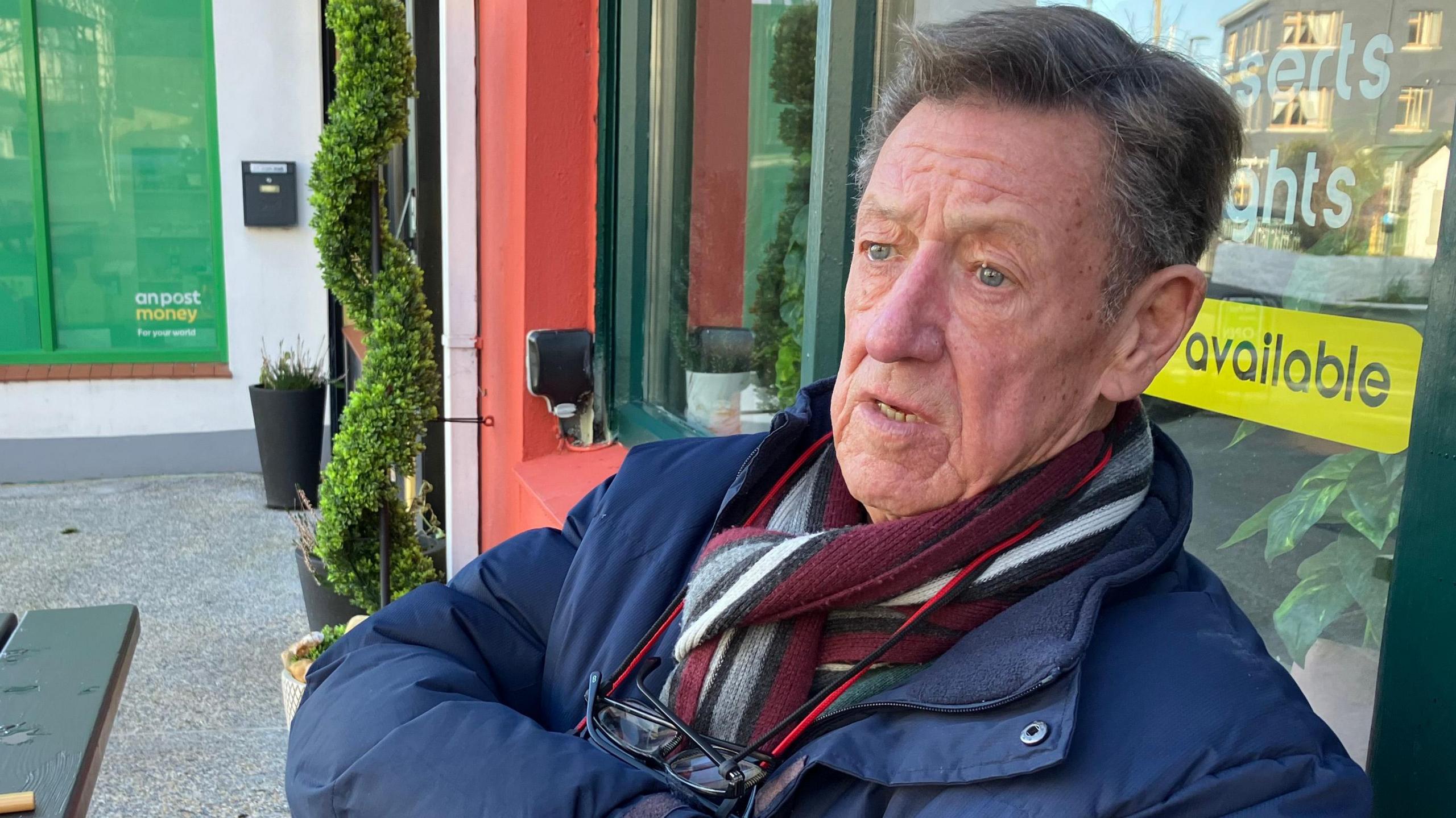 William Lane, who has grey hair, is wearing a dark blue jacket and red scarf. He has glasses handing from his neck and is sitting at tables outside a cafe. 