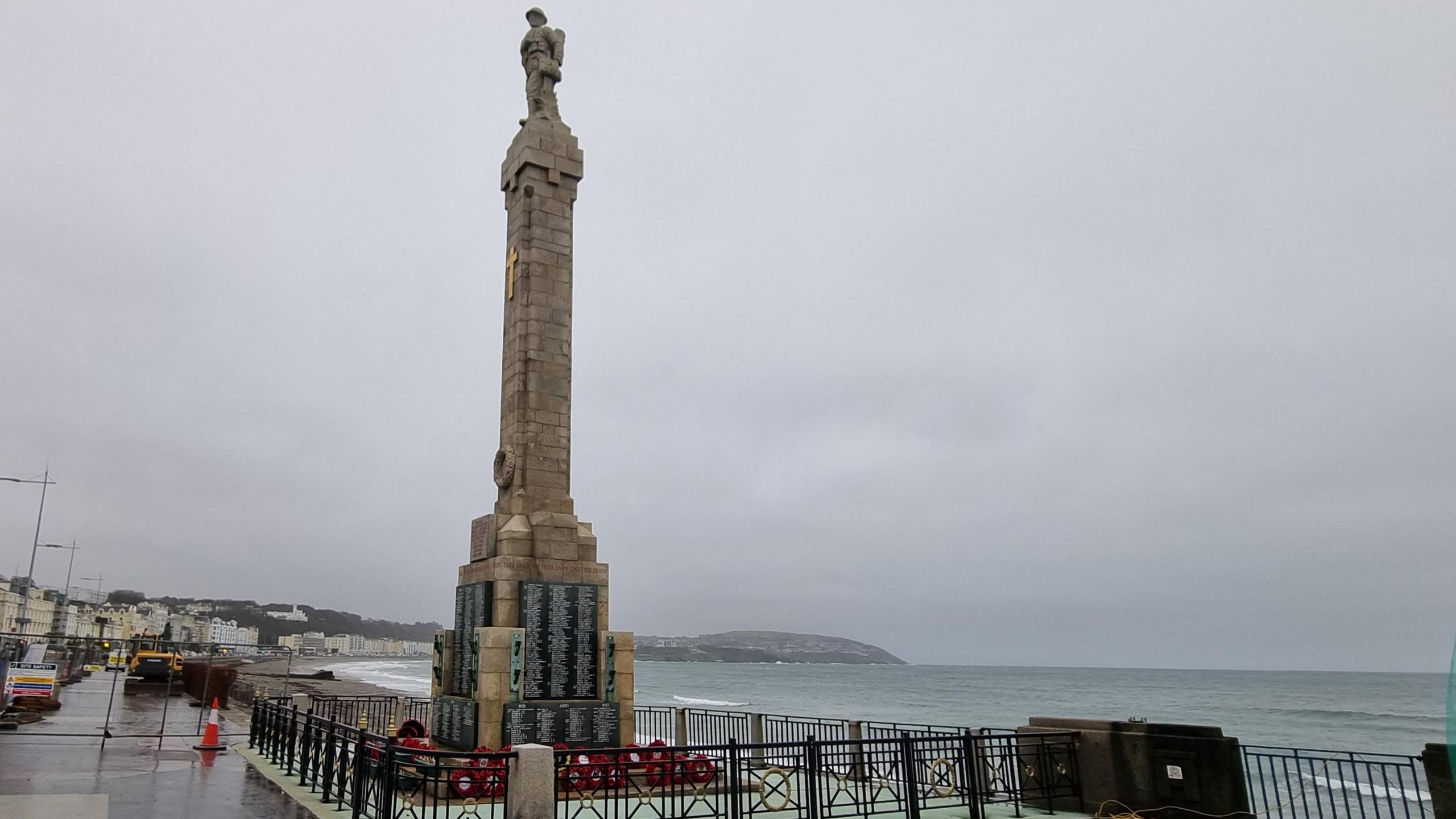 The war memorial on Douglas Promenade, which is a tall beige pillar with an infantry soldier on top. There is a gold cross on the memorial and poppy wreaths surrounding it.