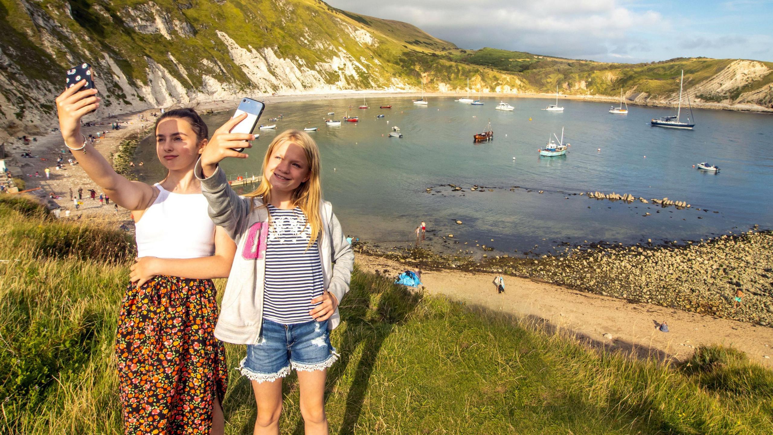 Two young girls hold up mobile phones on a grassy bank above Lulworth Cove, which is filled with small boats on a sunny day.