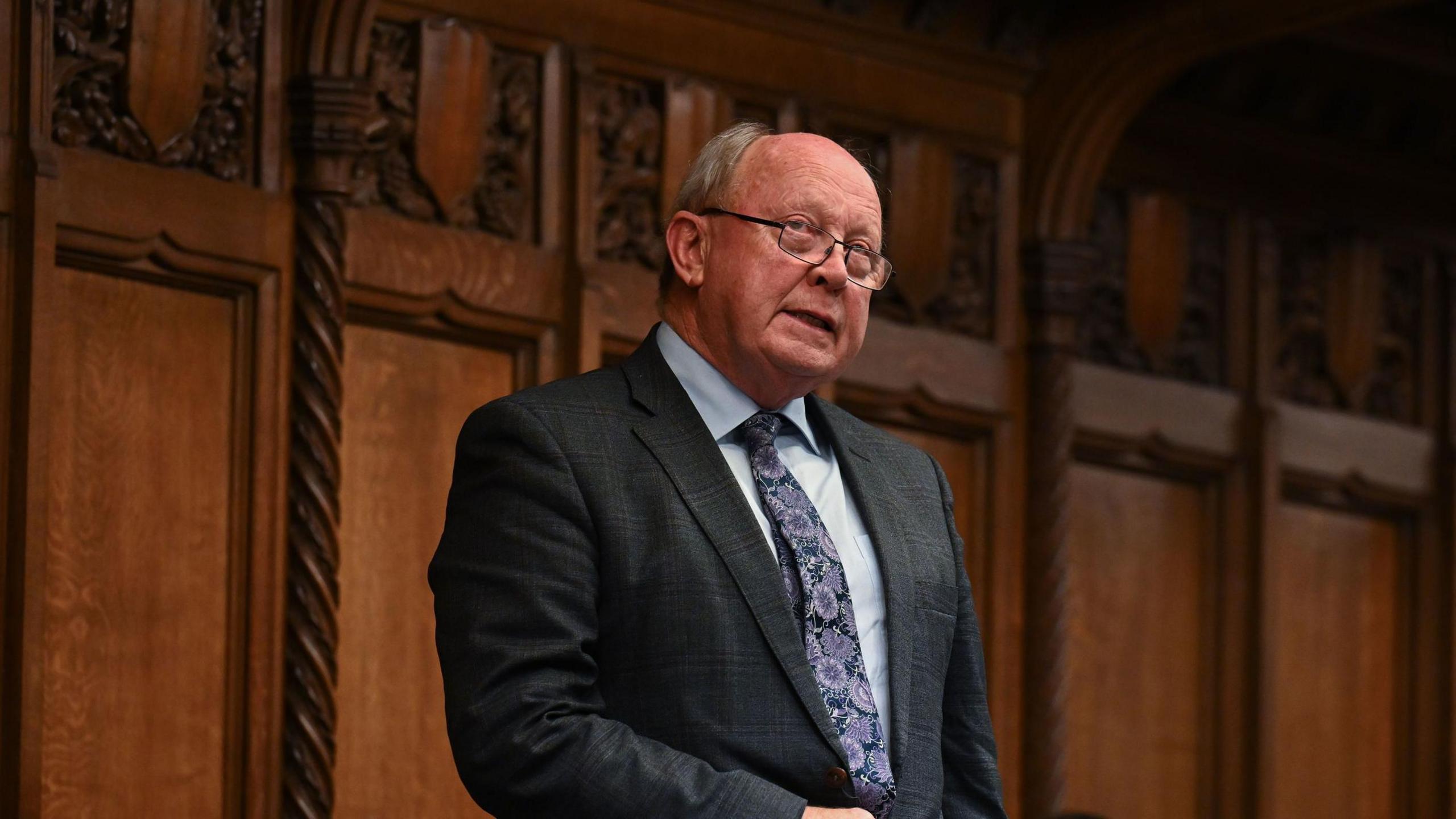 Jim Allister is bald on top with light hair at the sides. He is speaking in Parliament as he wears a dark suit, light blue shirt and patterened tie