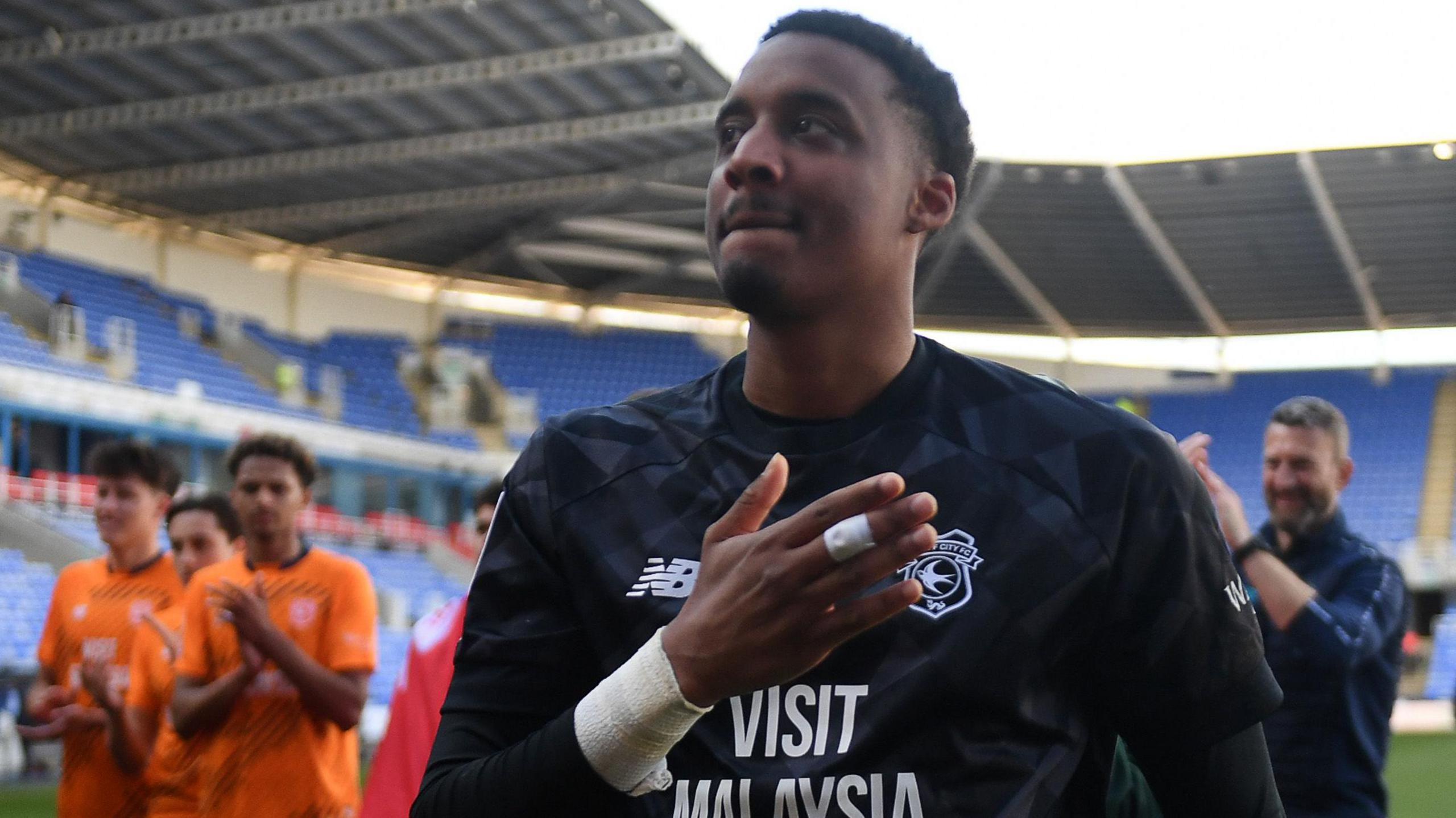 Nathan Trott in black Cardiff goalkeeper shirt, with his raised right hand partially over the club badge as figures including players behind him applaud