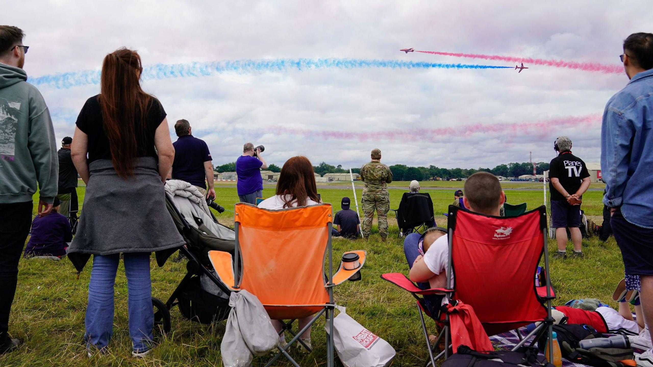 People are sitting on camping chairs and standing on grass watching a flight display. The aircraft are colouring the sky in red and blue.