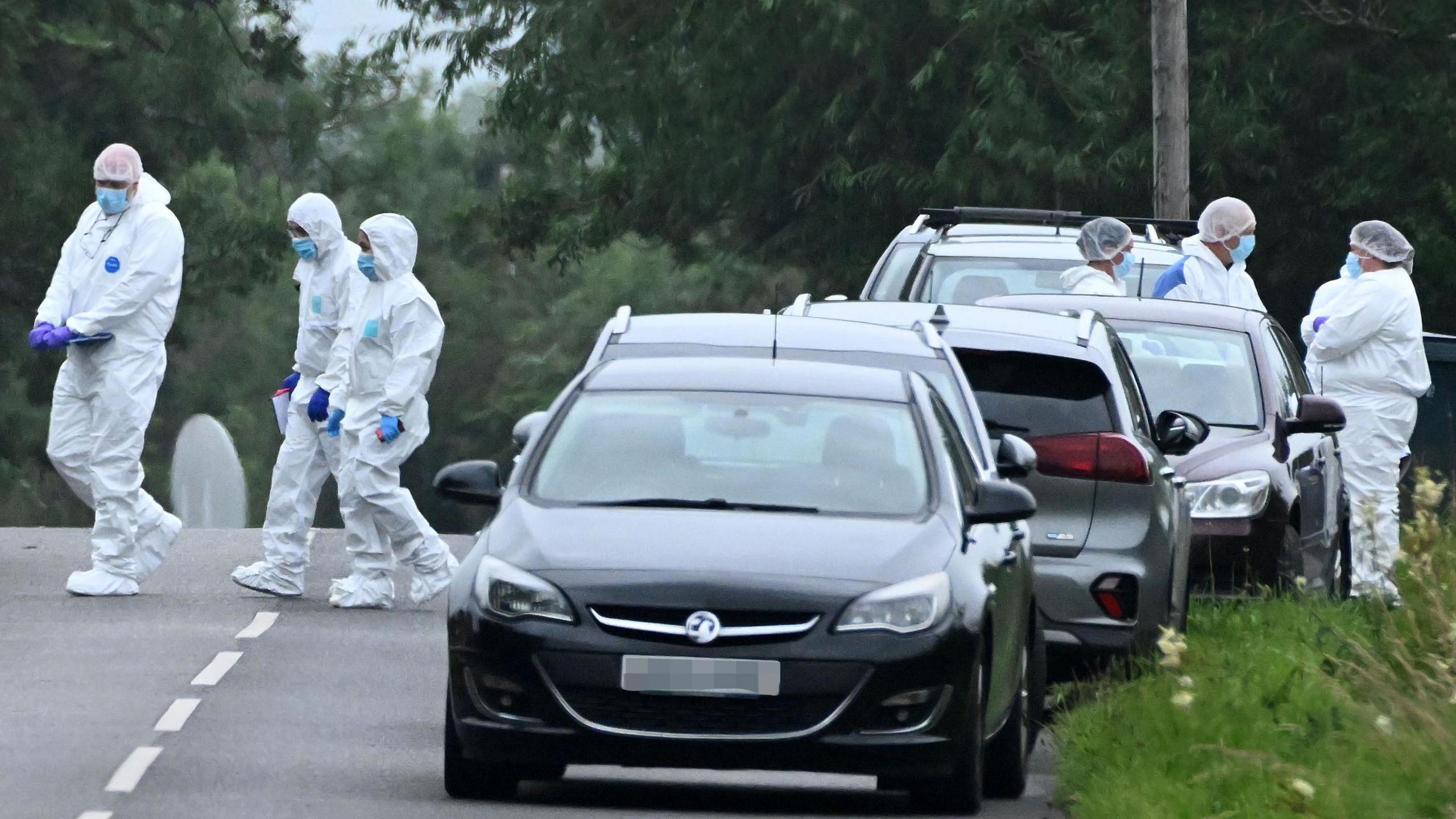 A number of people dress in white crime scene suits are pictured standing on a road beside a uniformed police officer. They all have their faces covered with blue masks. Beside them is a line of grey cars.