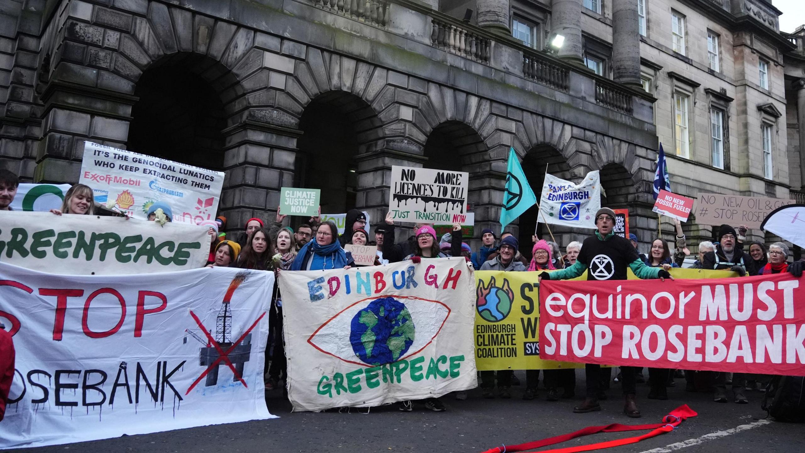 A group of protesters from climate organisations outside the Court of Session in Edinburgh. From left to right, one group are holding a greenpeace banner, which has the name of the organisation in green on a white banner. Below that is the words "Stop Rosebank" with stop in red and Rosebank in black. An image of an oil platform with a red X over the top is next to it. The next banner reads "Edinburgh" in multi-coloured letters and Greenpeace in green on a light-coloured banner either side of a globe in blue and green with a red eye shape surrounding it. The final banner is red with the words "equinor must stop Rosebank" in white letters.
