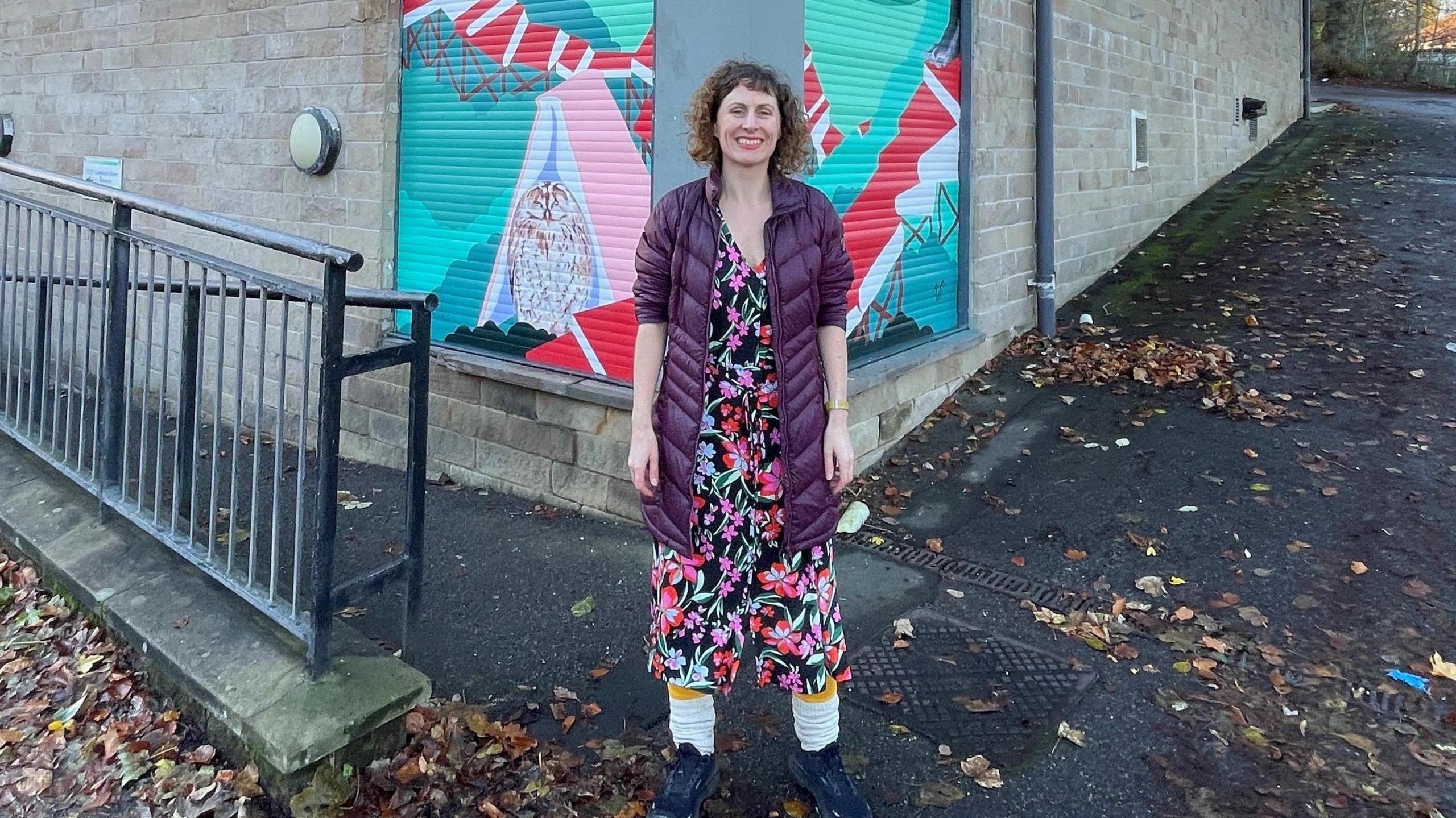 A woman is standing in front of a building smiling at the camera.  She is wearing a long purple coat with a bright black pink and red flowery dress underneath with white leg warmers and black trainers.