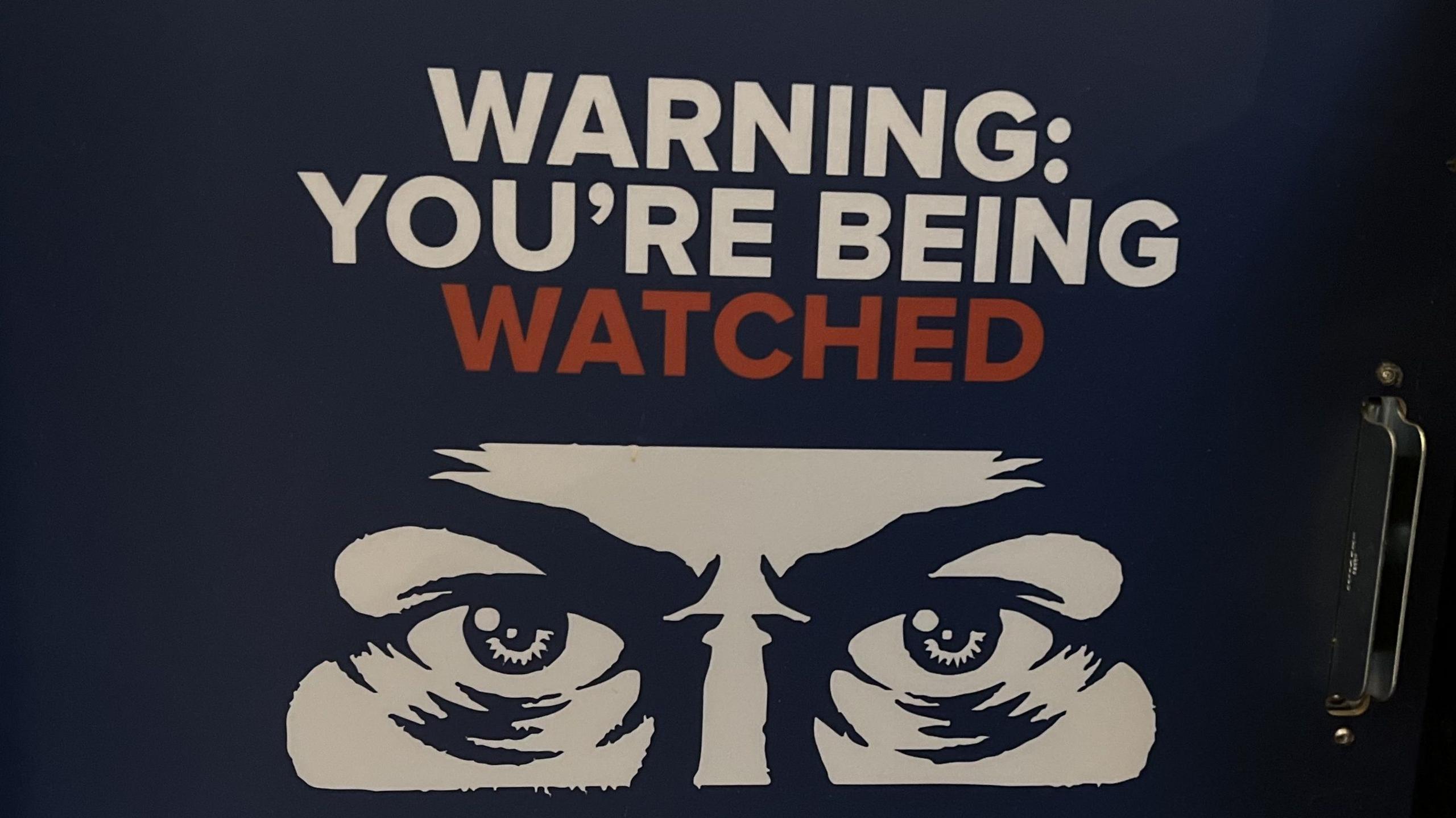 Sign with the words "warning you're being watched" above a drawing of eyes