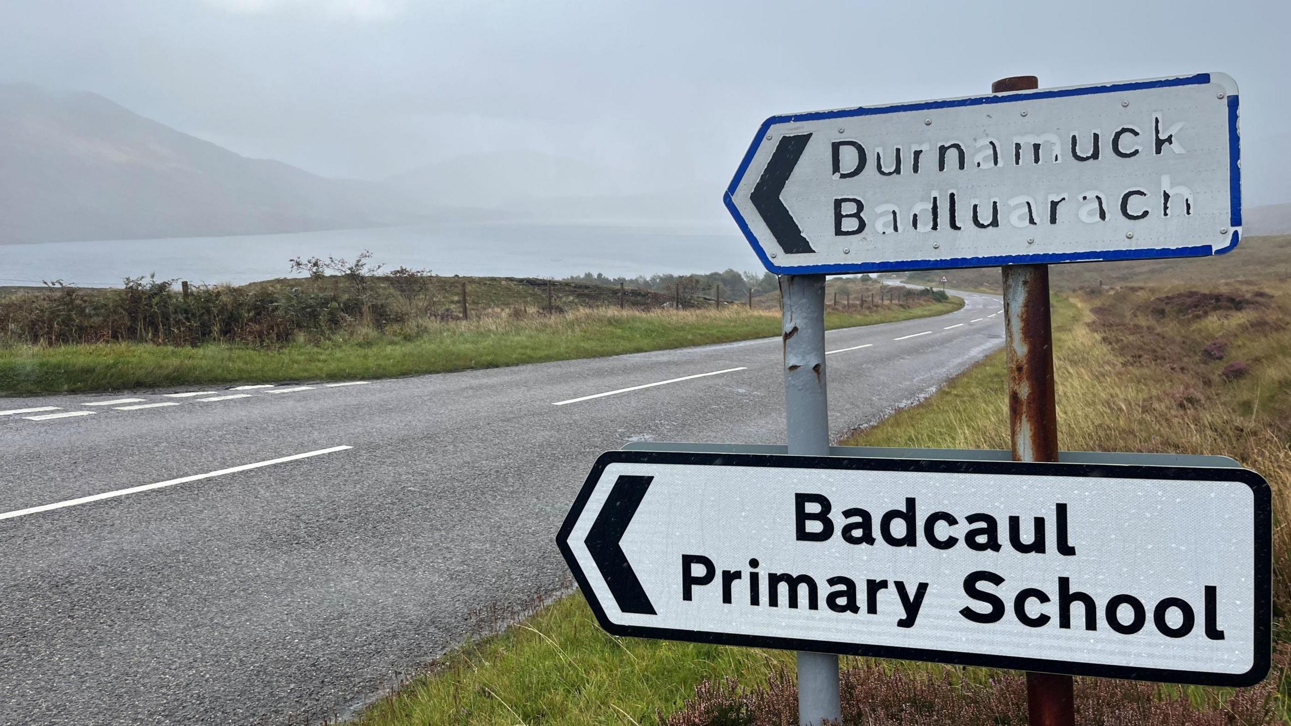 A sign pointing towards Badcaul Primary School. It has black writing on a white background and is in front of a road on a grass verge.