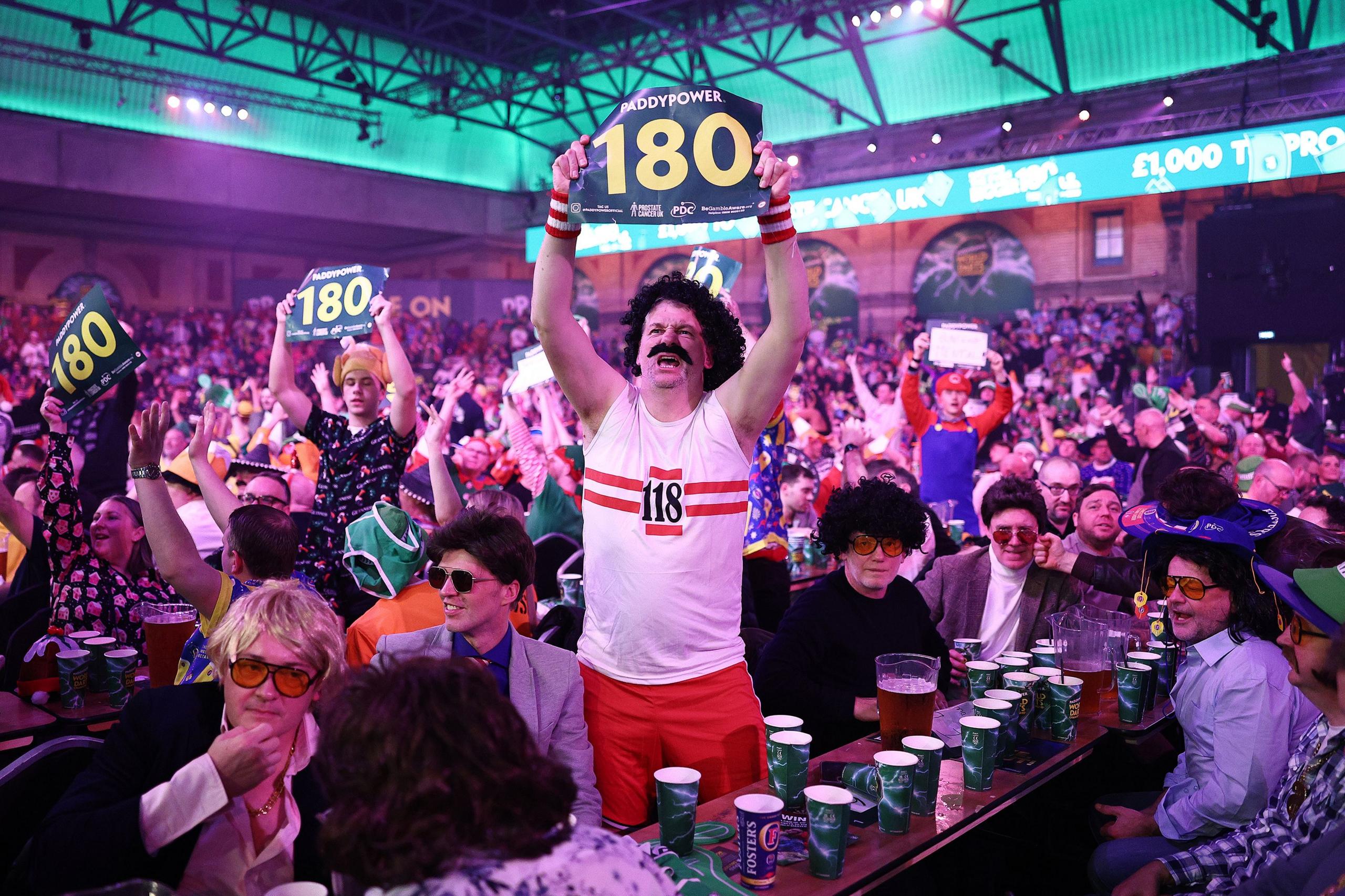 A lively crowd at an indoor darts event with bright green lighting and large banners overhead. In the foreground, a person dressed in a retro sports outfit with a white tank top and red shorts stands on a table holds a sign. The table is covered with numerous cups and pitchers of drinks. The background shows a packed audience cheering and waving signs, creating a festive atmosphere.
