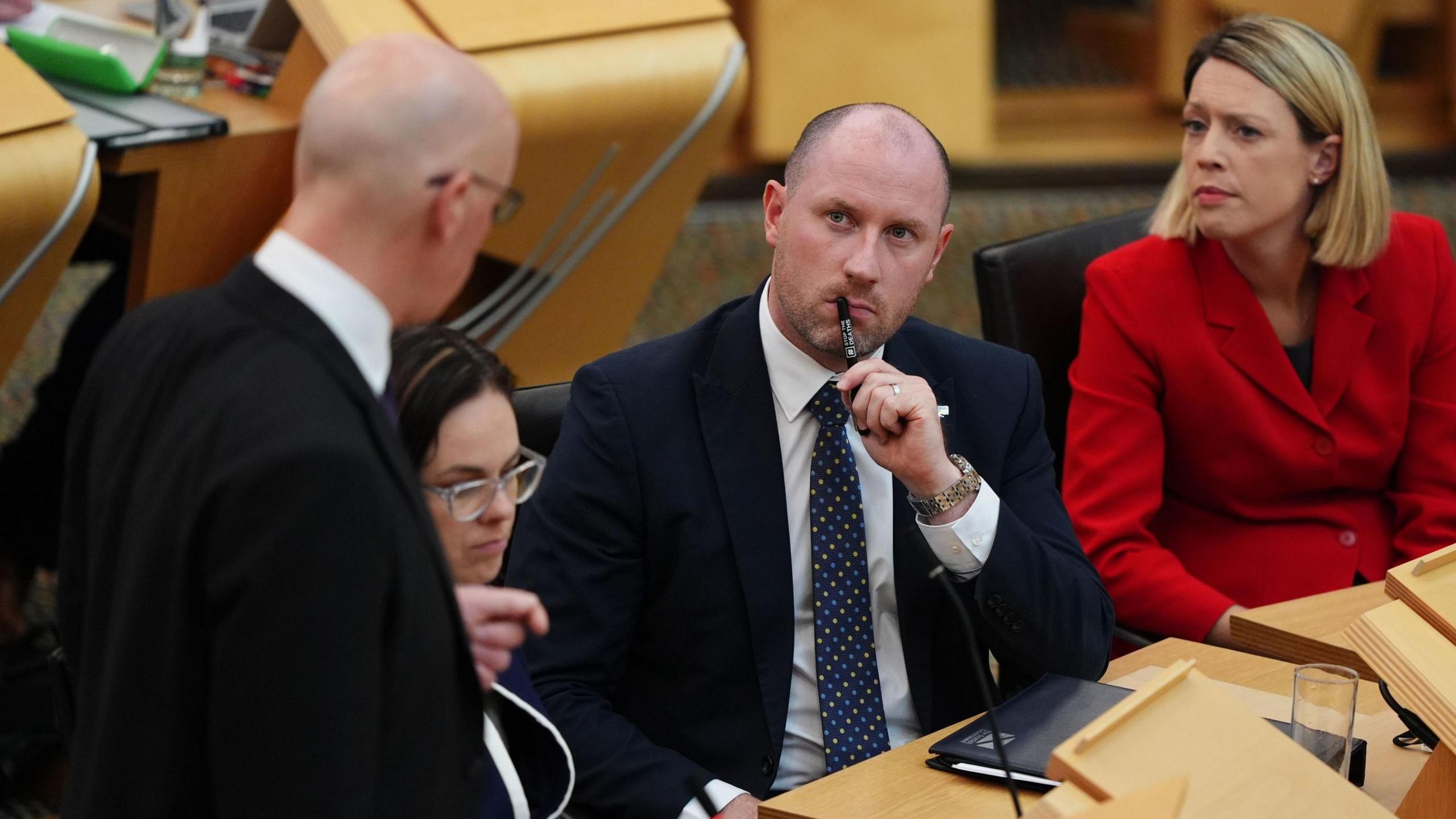 Neil Gray sitting inside the Scottish Parliament chamber with a pen in his mouth and a folder on the desk. He is wearing a dark suit and tie and is listening to John Swinney who has his back to the camera. Jenny Gilruth is sitting next to Gray wearing a bright red jacket and Kate Forbes sits between Swinney and Gray.