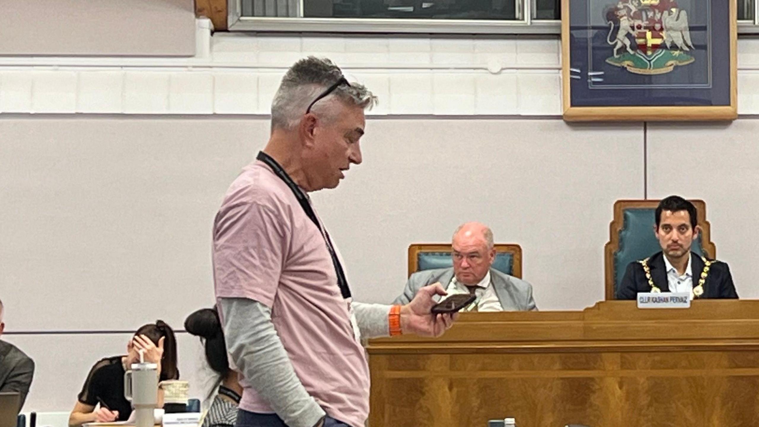 Councillor Matt Dimond-Brown, wearing a pink top and with glasses perched on his head, looks down at his phone while standing in a council chamber presenting a motion.