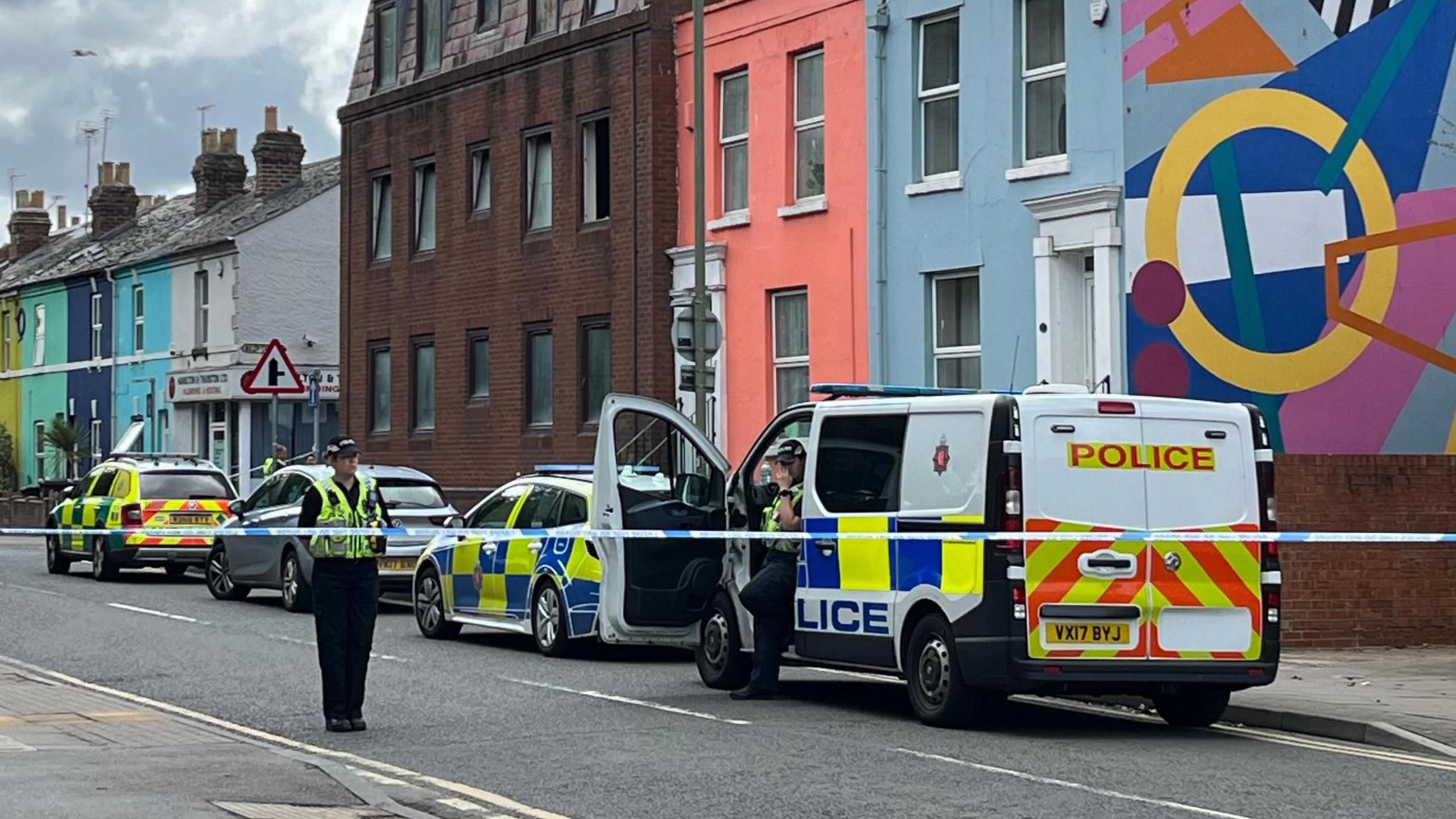 Police tape cordons off a road in Gloucester, which has colourfully painted houses dotted along it. There are a number of police vehicles - including a van - parked along it. A police officer is stood behind the cordon, surveying the surroundings, while another climbs out of the passenger door of the van.