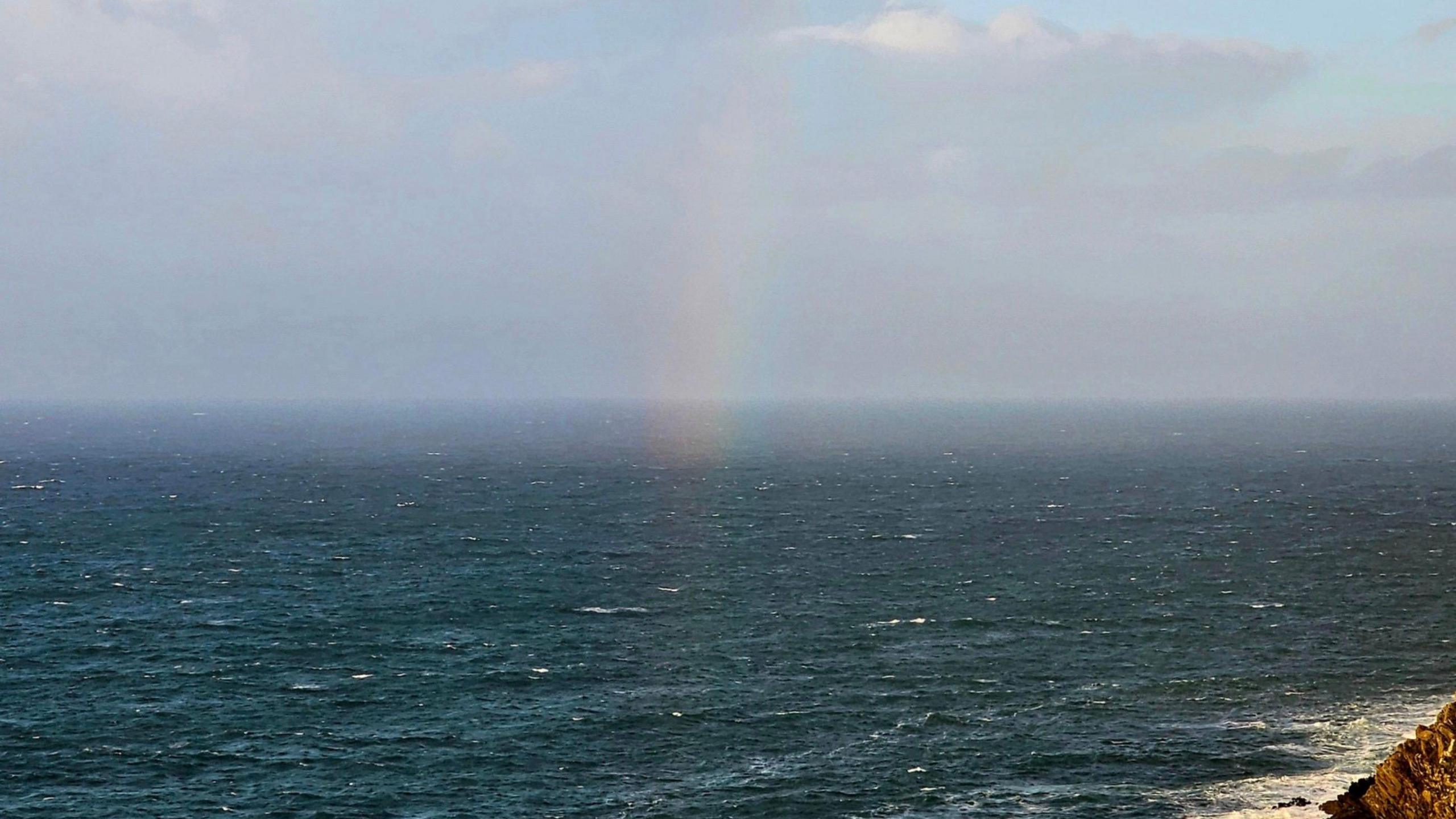 A picture taken off of the coast of Polperro. To the bottom right of the image is a rock. There is also a rainbow pictured in the centre of the image above the body of water.