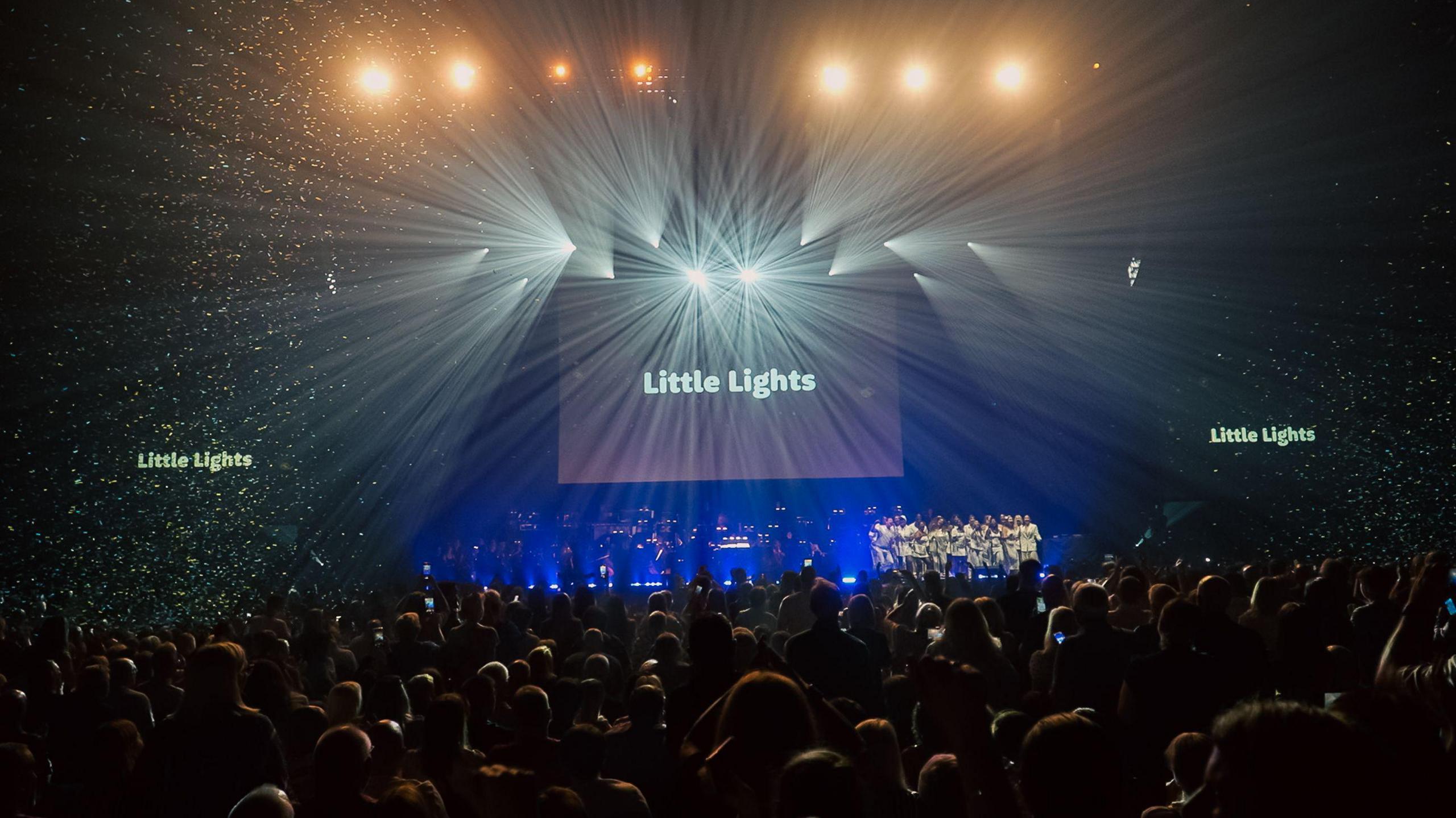 Thousands of people pack an arena as performers are on stage and the words 'Little Lights' appear on three big screens.