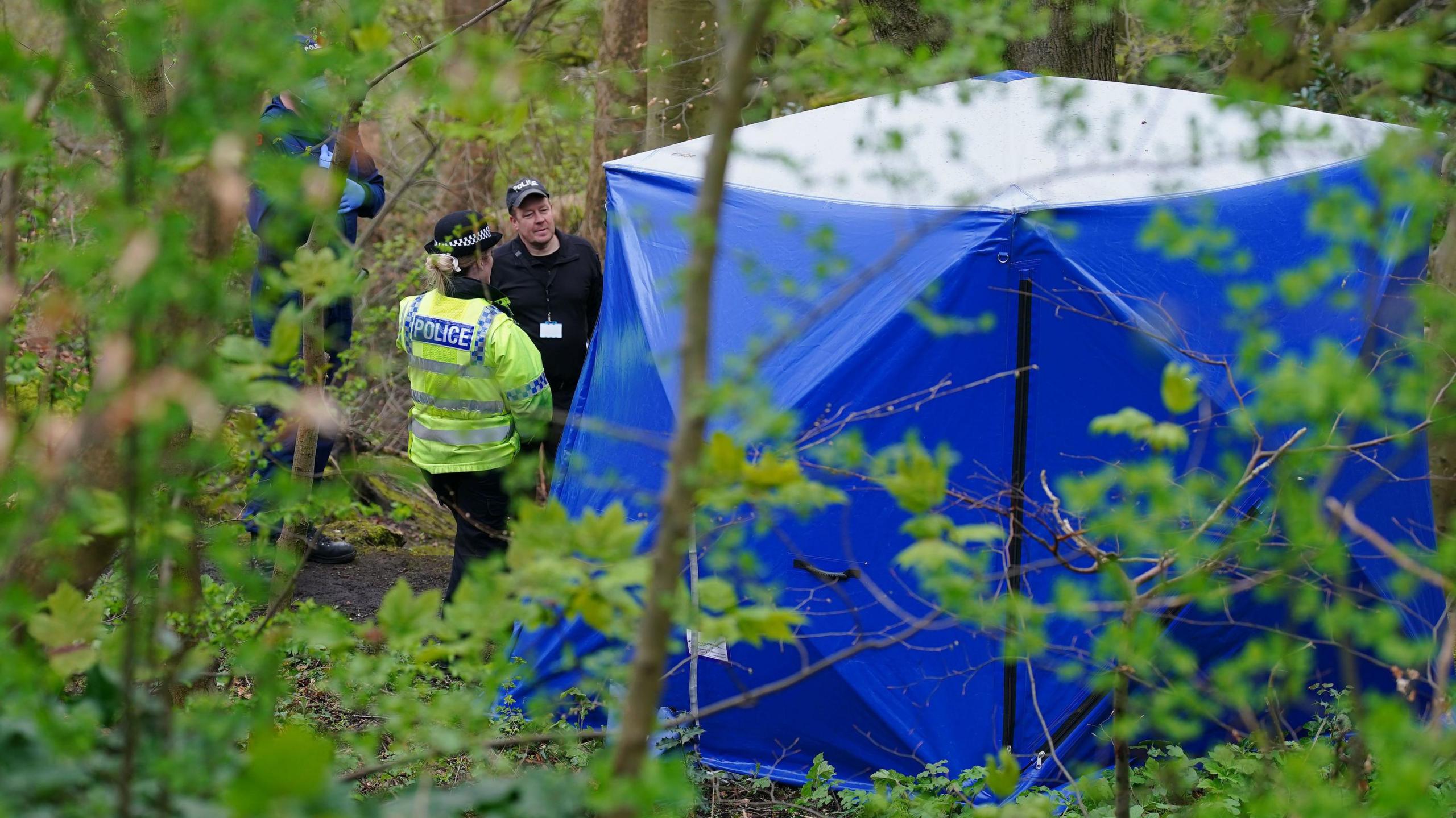 A blue tarpaulin forensic tent is erected in the middle of a densely wooded area in Kersal Dale, Salford. A police officer in a hi vis jacket and a hat stands with her back to the camera. The back of her jacket reads 'POLICE' in blue and white lettering. Opposite her a man with a cap that reads 'POLICE' is stood talking to her. He is dressed all in black. Another officer, possibly in a blue forensic suit stands behind some trees close to them.