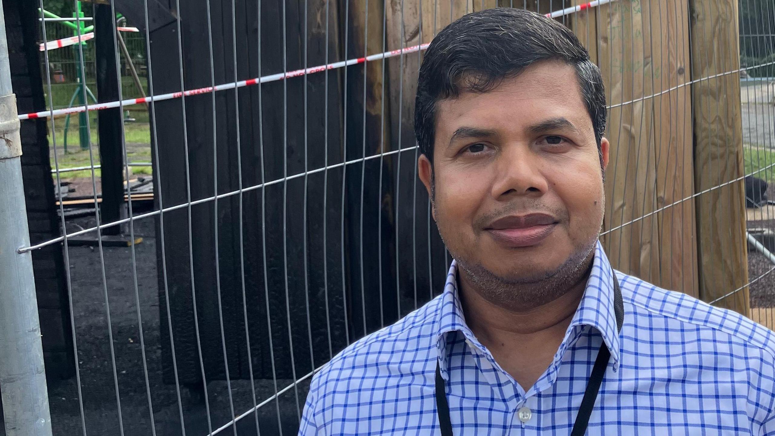 The head and shoulders of Councillor Delowar Hossain standing in front of metal fencing. Behind that is a view into the charred remains of the playground castle.