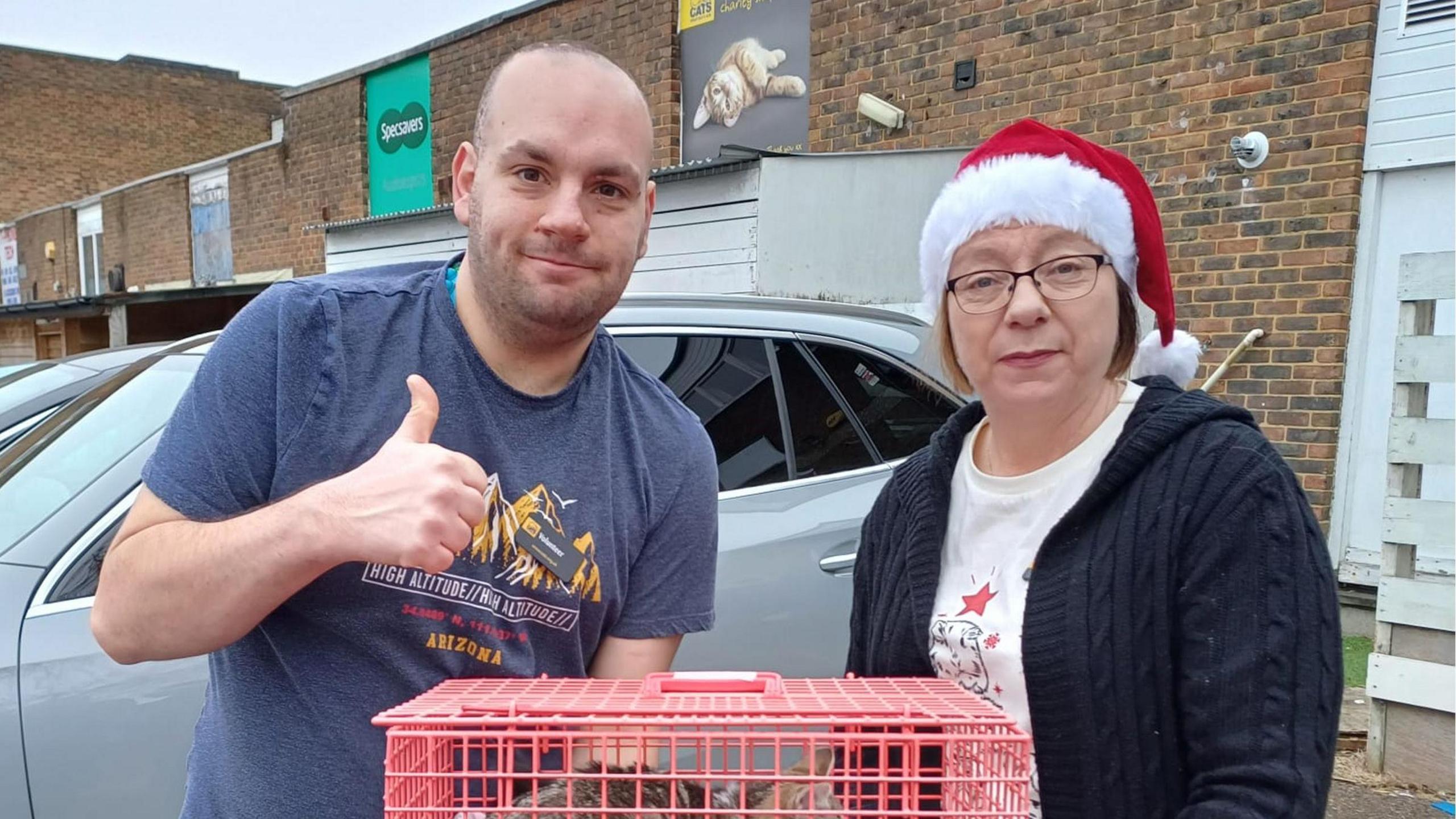 Mel Gunn is wearing a red santa hat, white top and navy jumper whilst holding a red crate. Also holding the crate is a bald man wearing a blue t-shirt.