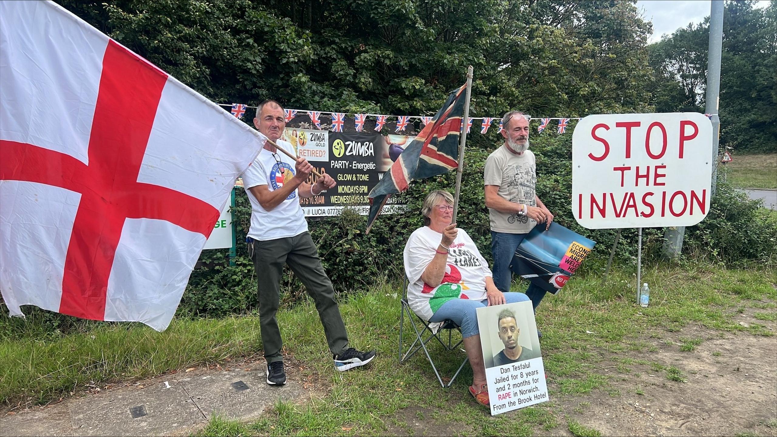 Picture shows three men, one holding a St George Flag and the other is sitting down while holding a poster. The third man is standing next to a sign that reads 'Stop the invasion'.