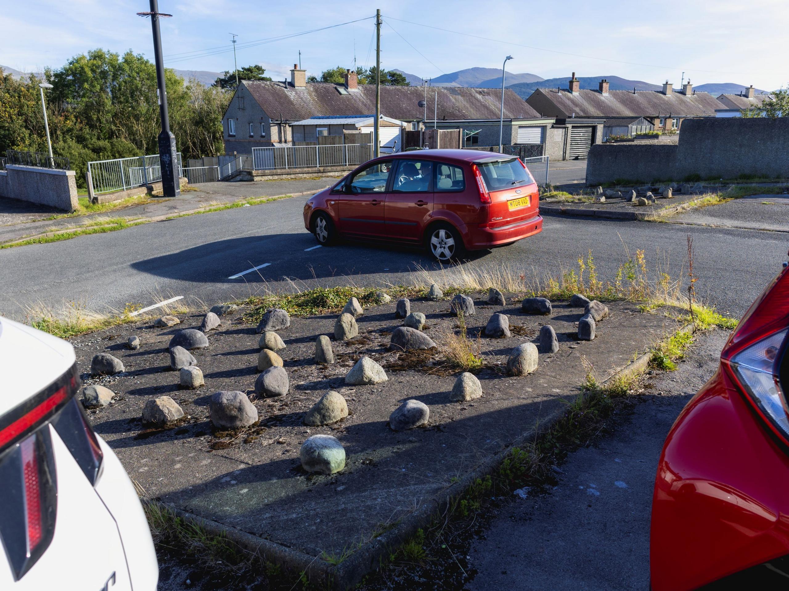 A red car is pulling out onto a road at a junction with a row of garages beyond. Behind it is a square of concrete with big stones arranged on top of it, and a glimpse of two cars, one red, one white, parked behind.