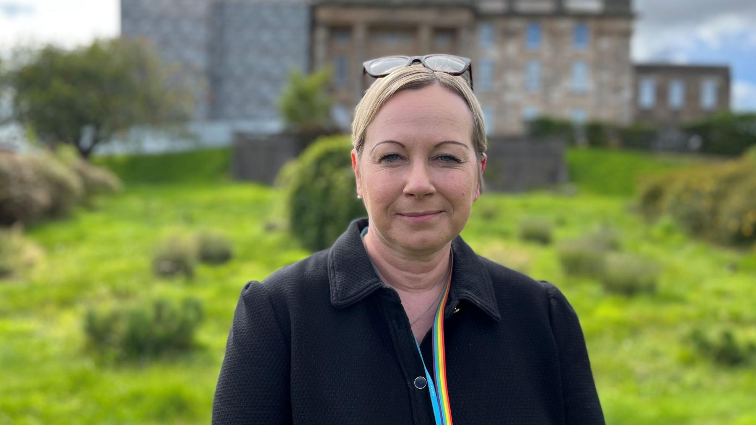 NHS Grampian chief executive Laura Skaife-Knight is standing outside. Dr Gray's hospital is in the background. Skaife-Knight has blonde hair tied up and glasses on her head. She is wearing a black coat and multi-coloured lanyard.