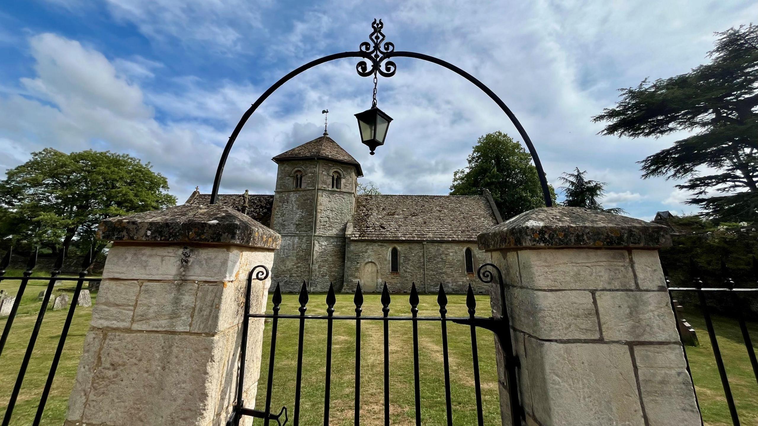An historic church called St Nicholas Church in the village of Ozleworth in the Cotswolds is pictured on a sunny day through its own gate, which has an old-fashioned lantern above it