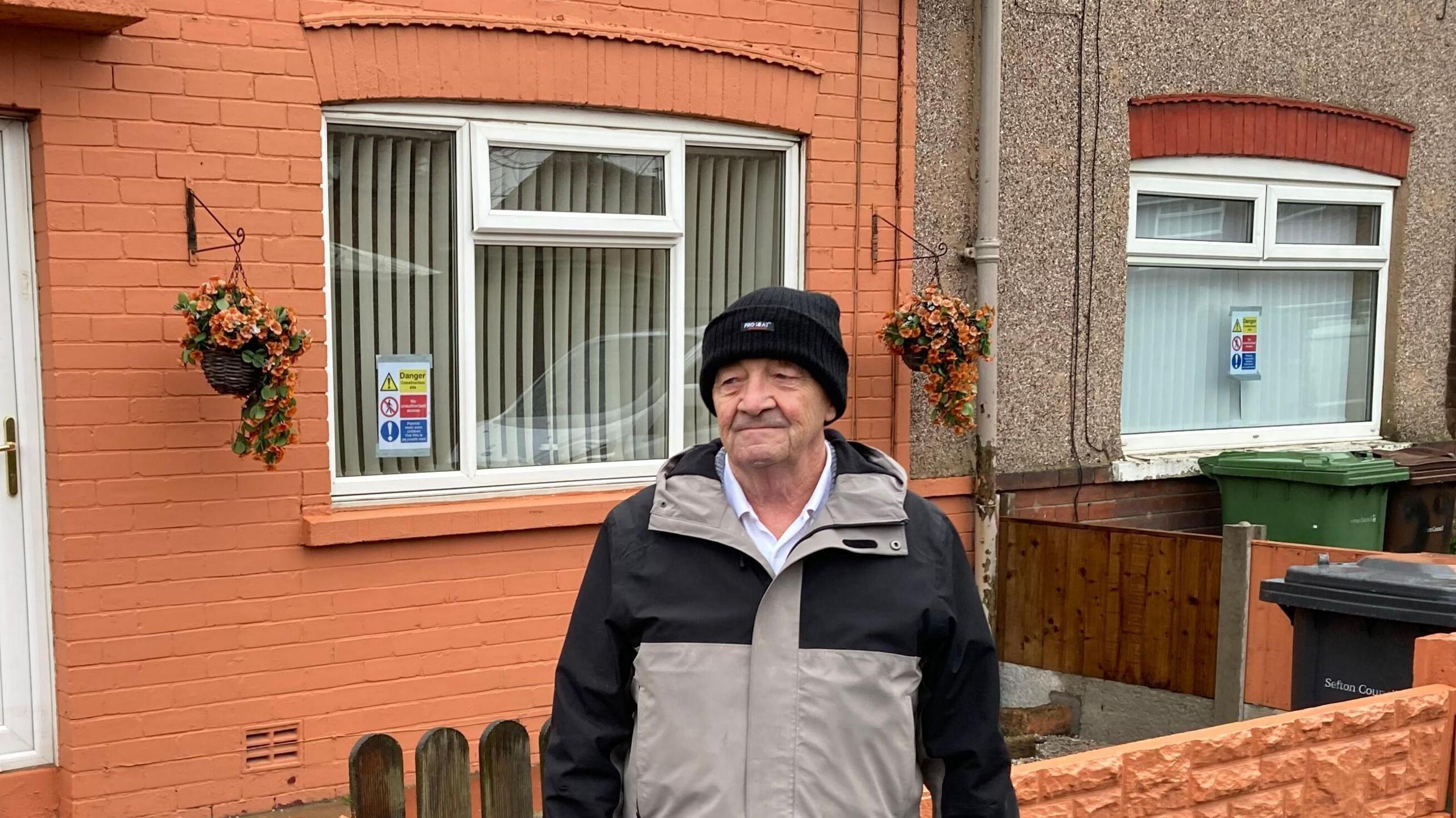 Sid Traynor, 80, wearing a black woollen hat, a beige and black overcoat and blue jeans, stands outside a terraced house which has an orange painted garden wall with a wooden gate