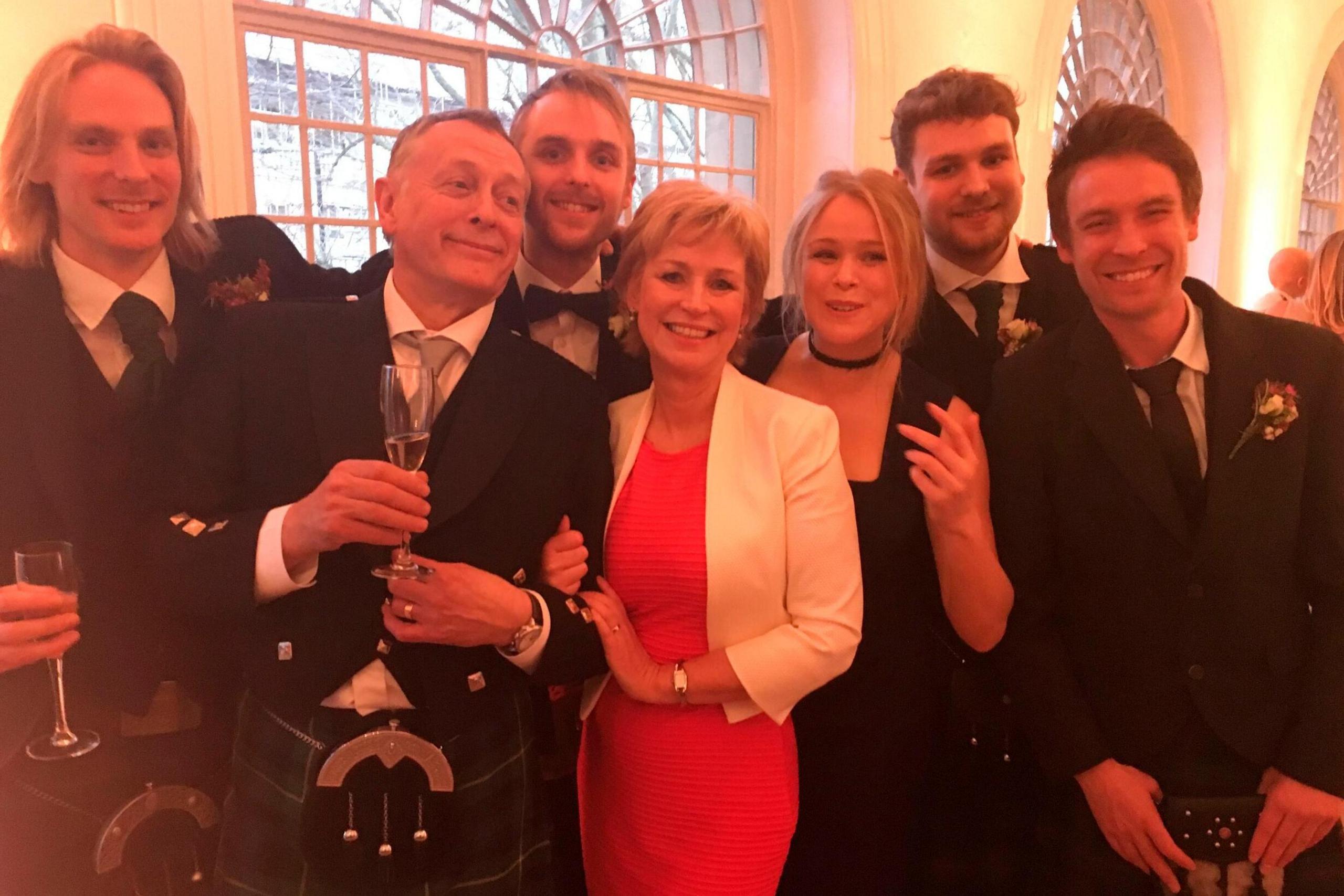 Sally Magnusson with her family in formal wedding clothes. Sally is in the centre wearing a red dress and white jacket and her daughter Anna Lisa is on her right wearing a black dress. Sally's husband Norman is on her left in a kilt and sporran and formal wedding jacket. Also pictured are the couple's four sons Jamie, Rossie, Magnus & Siggy.