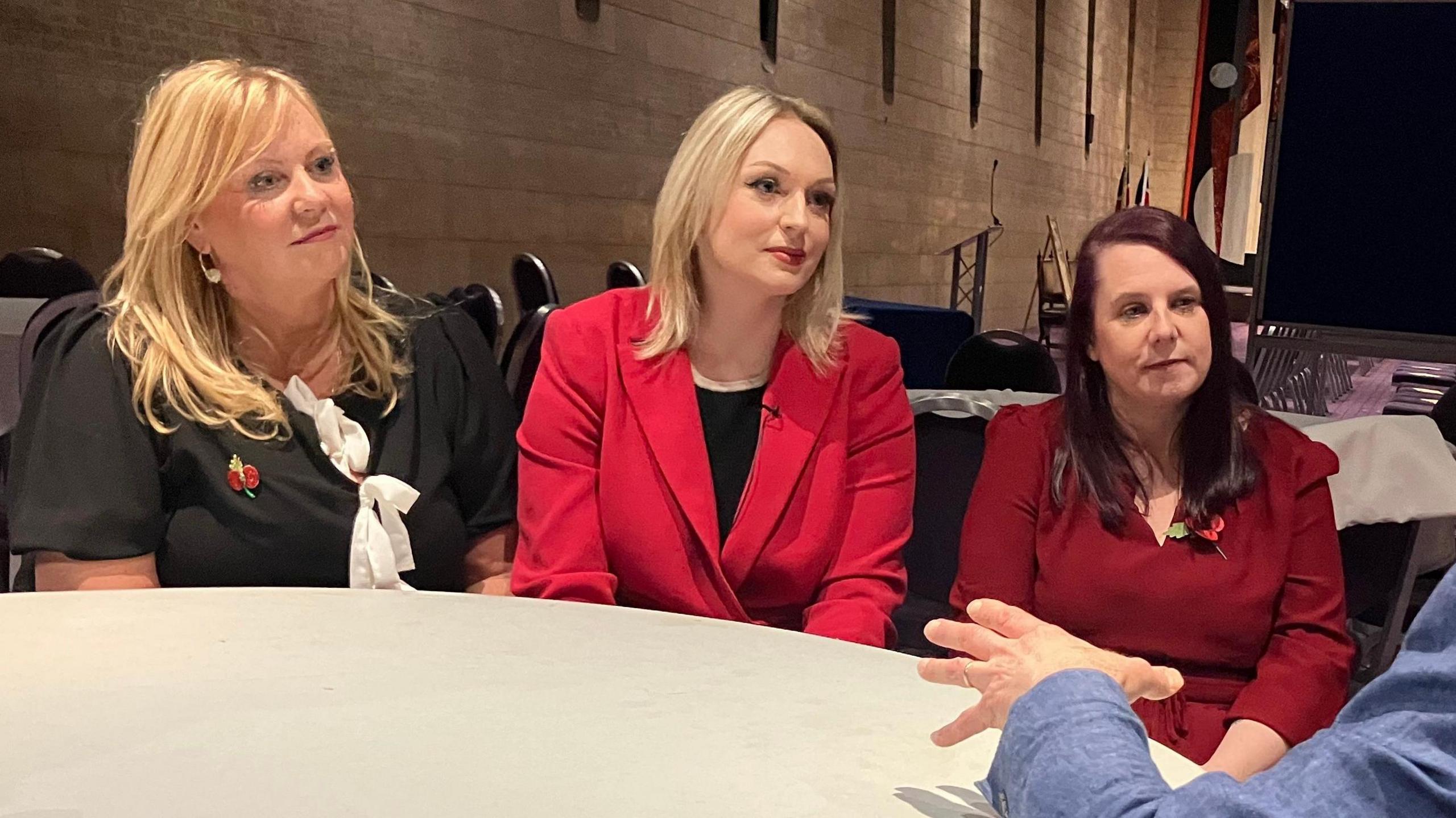 Nurses Lisa Lockey, Bethany Hutchison and Karen Danson sit at a table facing a reporter asking them questions. Lisa and Bethany both have long blonde hair and Karen has dark red hair. They are all smartly dressed with two of them displaying poppies.

