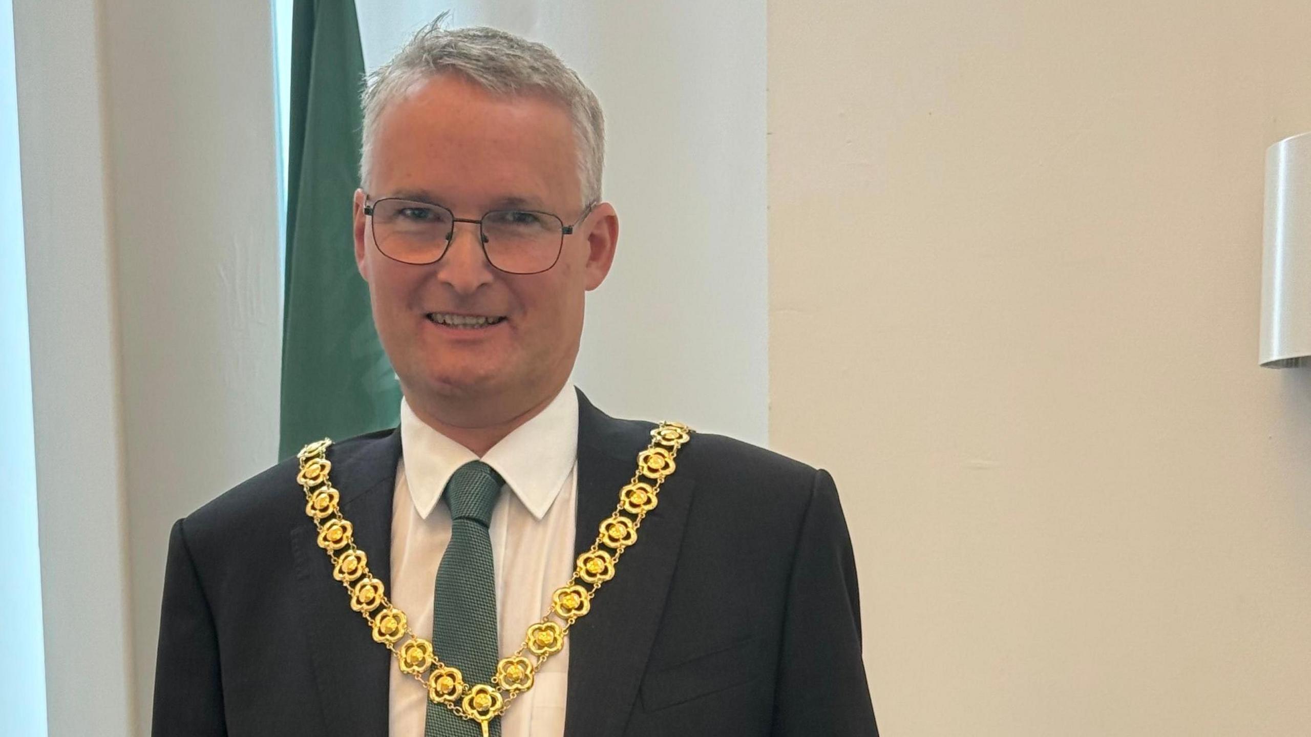 Councillor Louis Stephen wearing a mayoral chain inside the council building. He has grey hair and is wearing a dark suit, with white shirt and a green tie and wire-framed glasses.