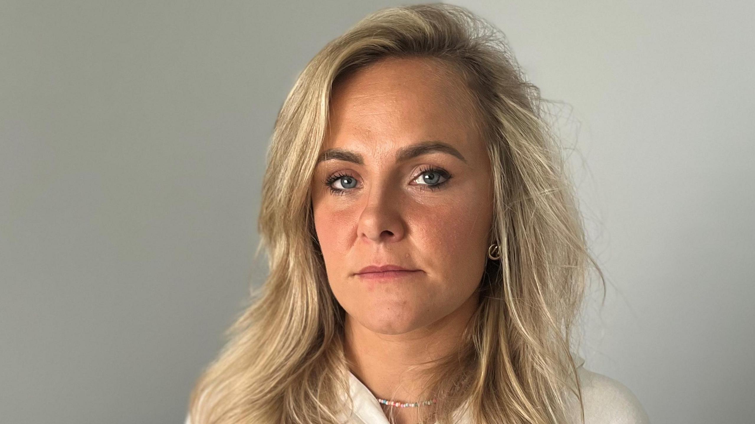 Hannah Farrell, a nurse, looks into the camera. She has a neutral expression on her face. We can see her from the shoulders up. She has shoulder-length blonde hair and is wearing a white or cream top. She has a metal chain around her neck. She is standing in a room, there is a plain grey-coloured wall behind her.