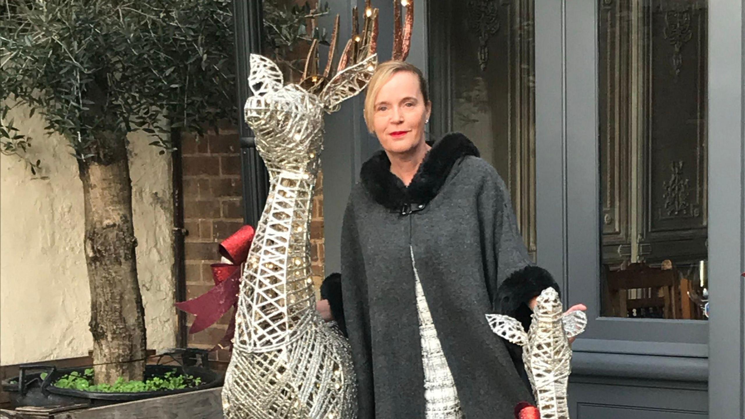 A woman wearing a dark grey cape over a white dress poses for a photograph while standing between two metal frame statues of deer.