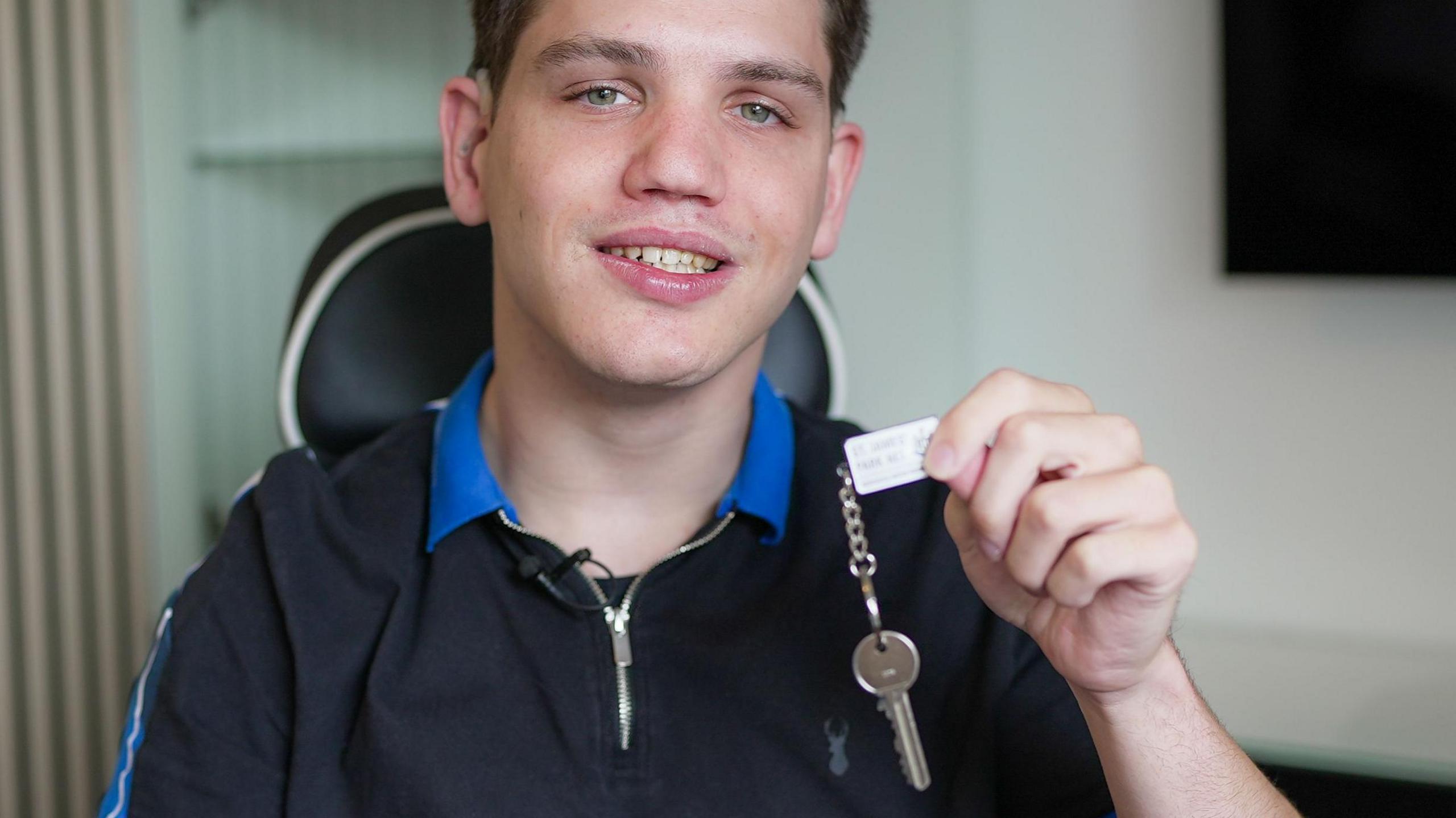 Elliot is sitting in his wheelchair and looking at the camera, holding up his new house keys in his left hand. He is smiling and wearing a black t shirt with a blue collar and a zip at the neck.