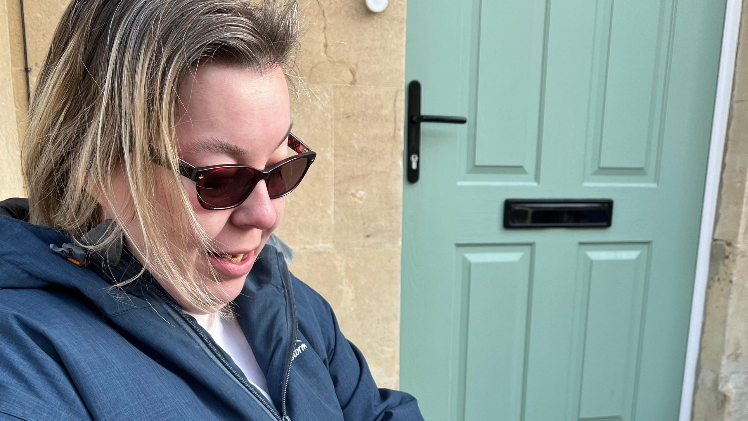 A blonde woman in sunglasses and a dark blue anorak and white T-shirt sits outside a house on the street where the Residents' Parking Zone is due to be brought in. 