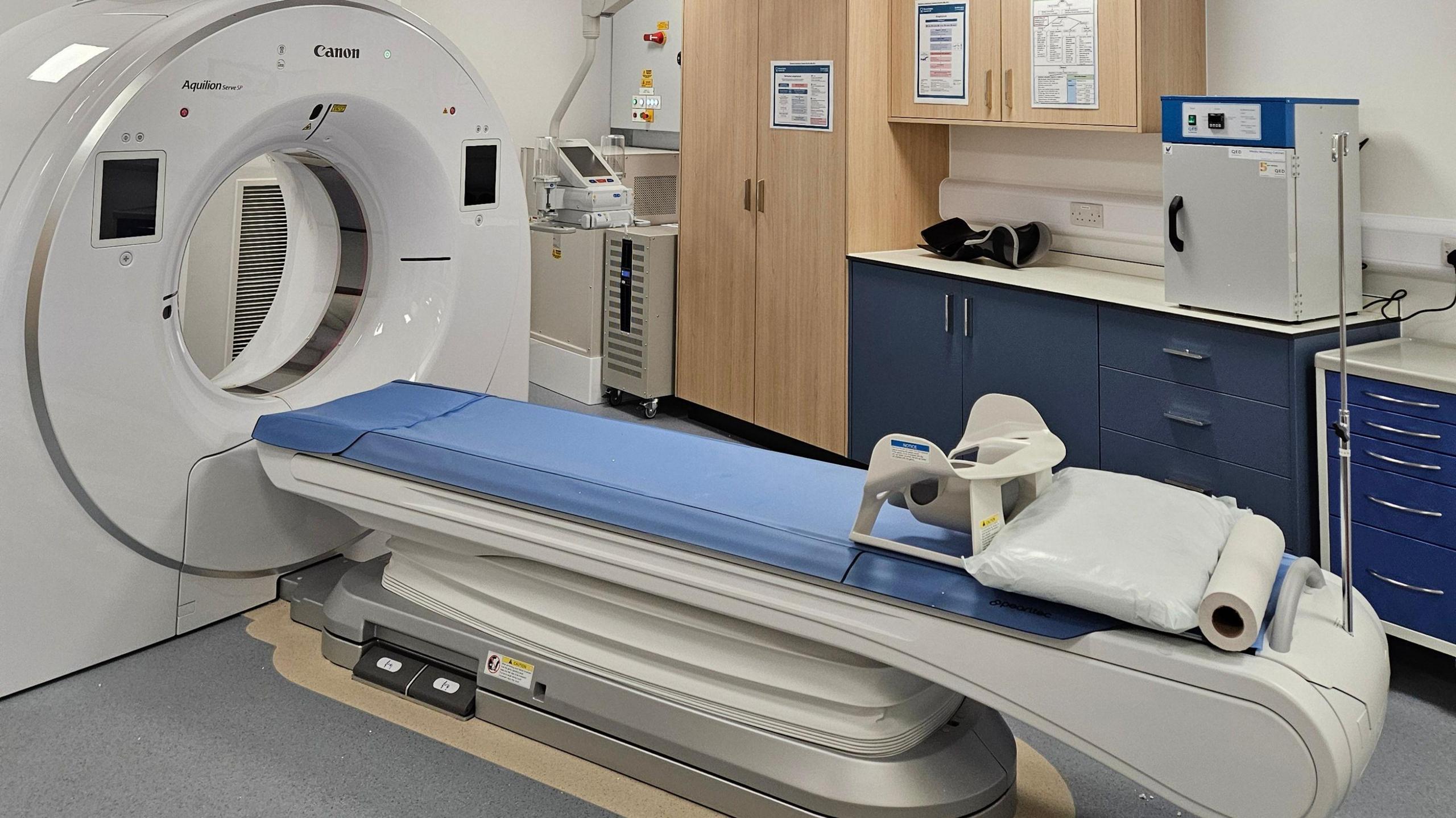 A large MRI scanner in a treatment centre room. A blue bed is in the centre of the room, attached to a large circular machine. There are blue cupboard doors around the edge of the room.