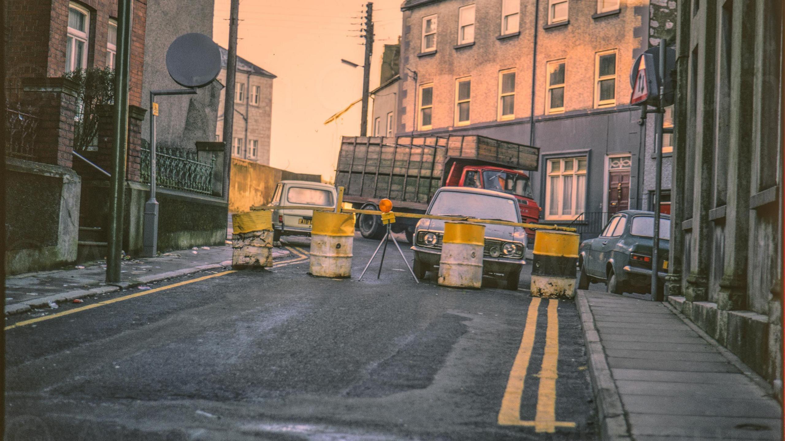 A digitalised photo from a film of oil drums acting as a barrier. Cars are parked on the far side. Abbey Street is blocked by a security barrier made from a series of concrete filled oil drums.