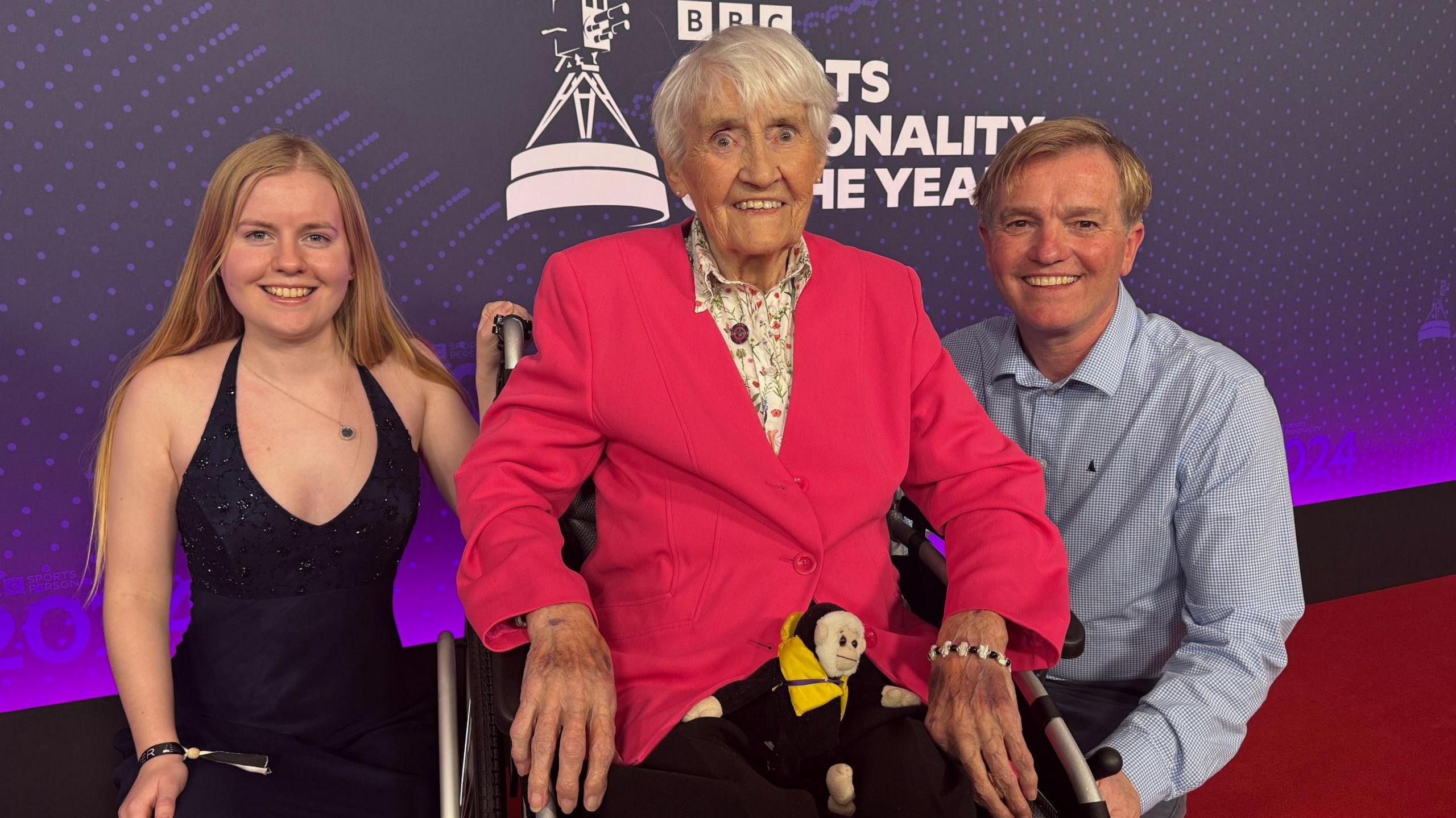 Jean Paton sits in her wheelchair looking directy at the camera, she has white, short hair and wears a bright fuschia pink blazer, buttoned up with a white collared shirt which has a green and pink floral print on it. She wears black trousers and has a toy monkey sitting in her lap, which has a yellow life jacket on. In the background is a purple sign reading 'BBC Sports Personality of the Year' and Ms Paton is on the red carpet for the event. To her left, is her granddaughter Alex, who has long blonde hair and wears a navy blue halterneck gown which has sequins on the top. Ms Paton's son, Rory, sits to her right, he has short blonde hair in a side parting and wears a blue and white checked shirt. They are all smiling at the camera.