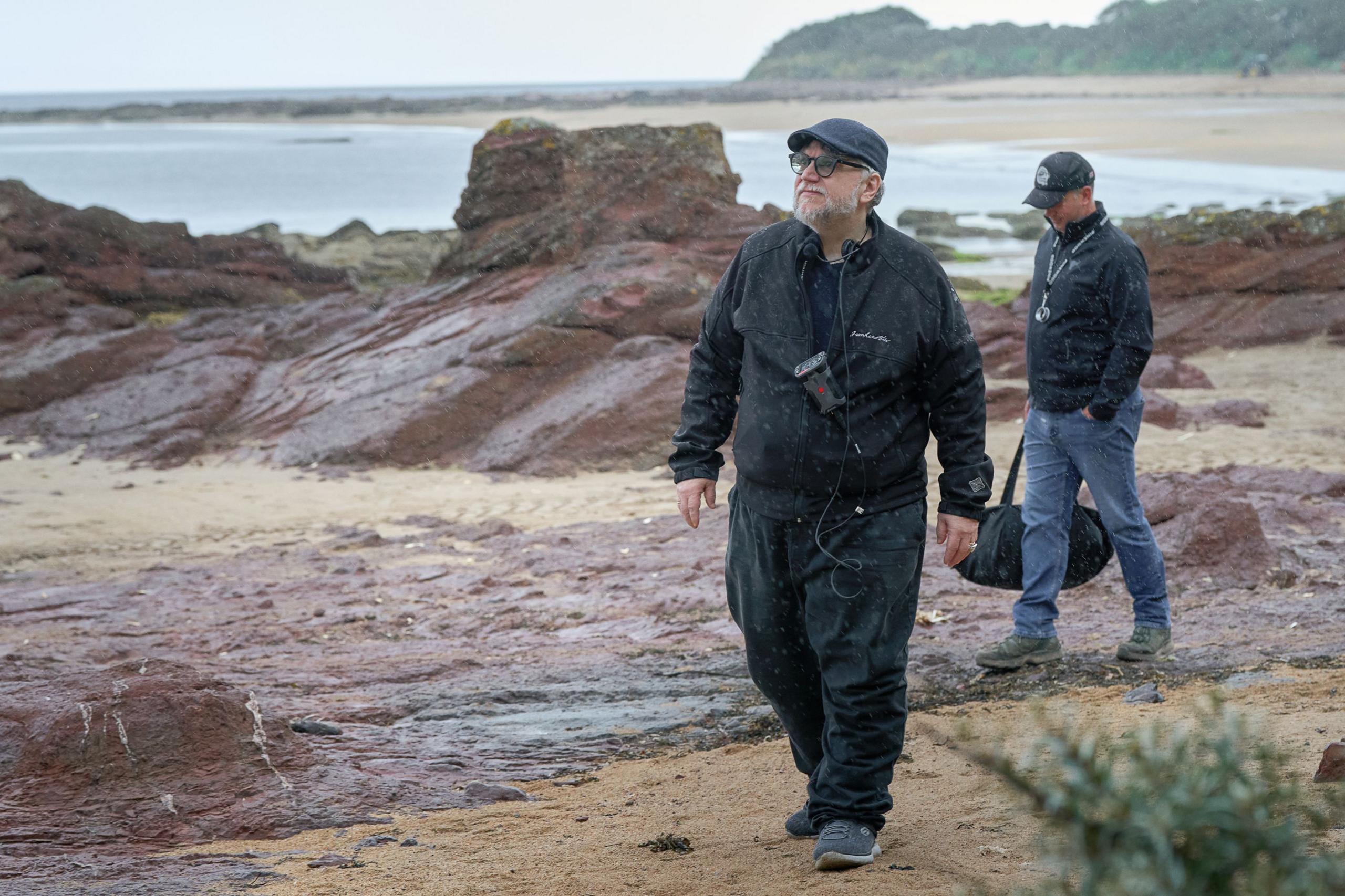 Guillermo Del Toro, in flat cap with all black outside wear, is walking along a beach staring out to sea. A man in jeans, baseball cap and carrying a bag, walks along behind him. There are red sandstone extrusions behind them as well as sea and a beach. It is very grey.