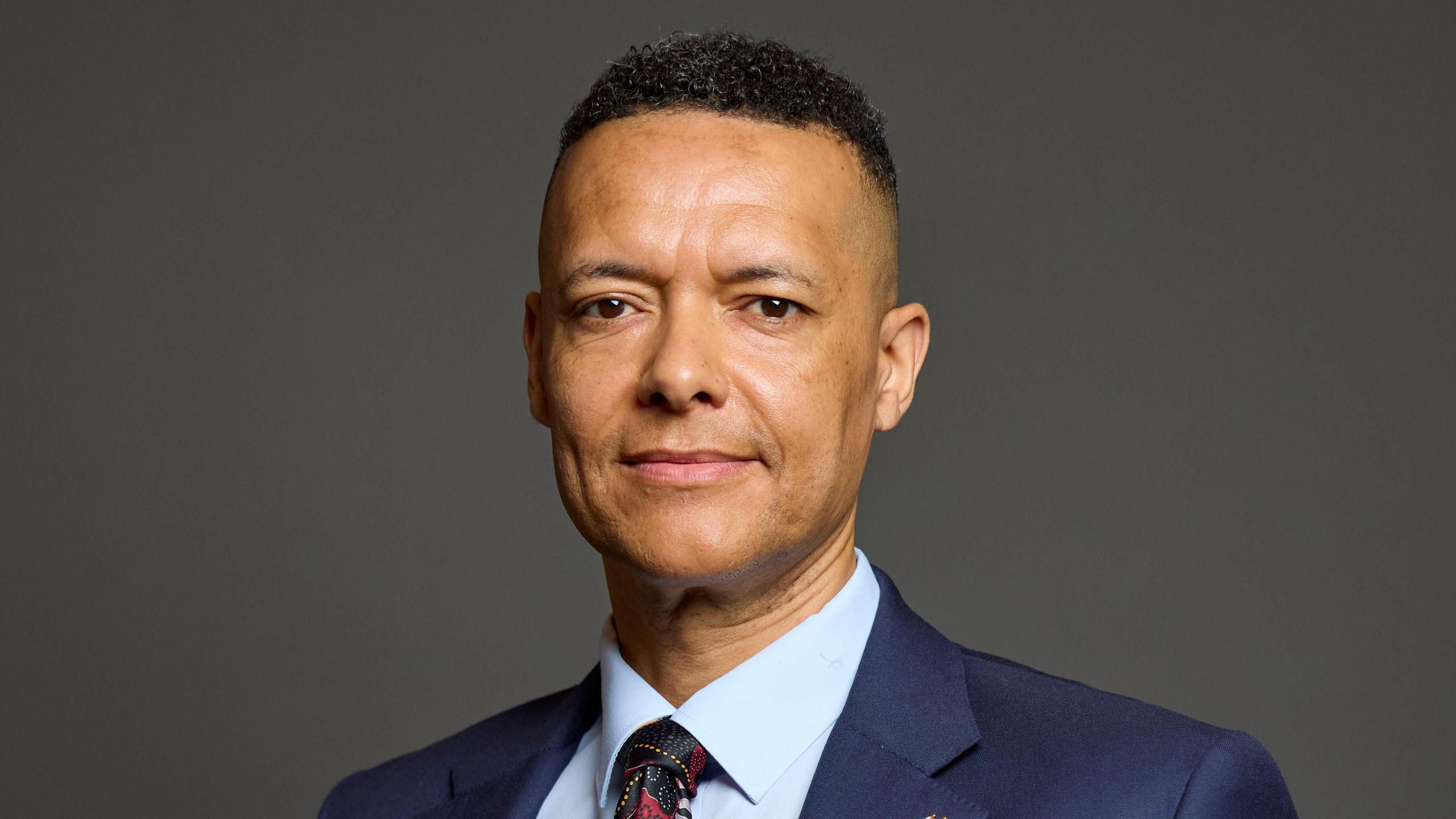 A smiling Clive Lewis looks directly at the camera, dressed in a suit and tie and standing against a dark background. 