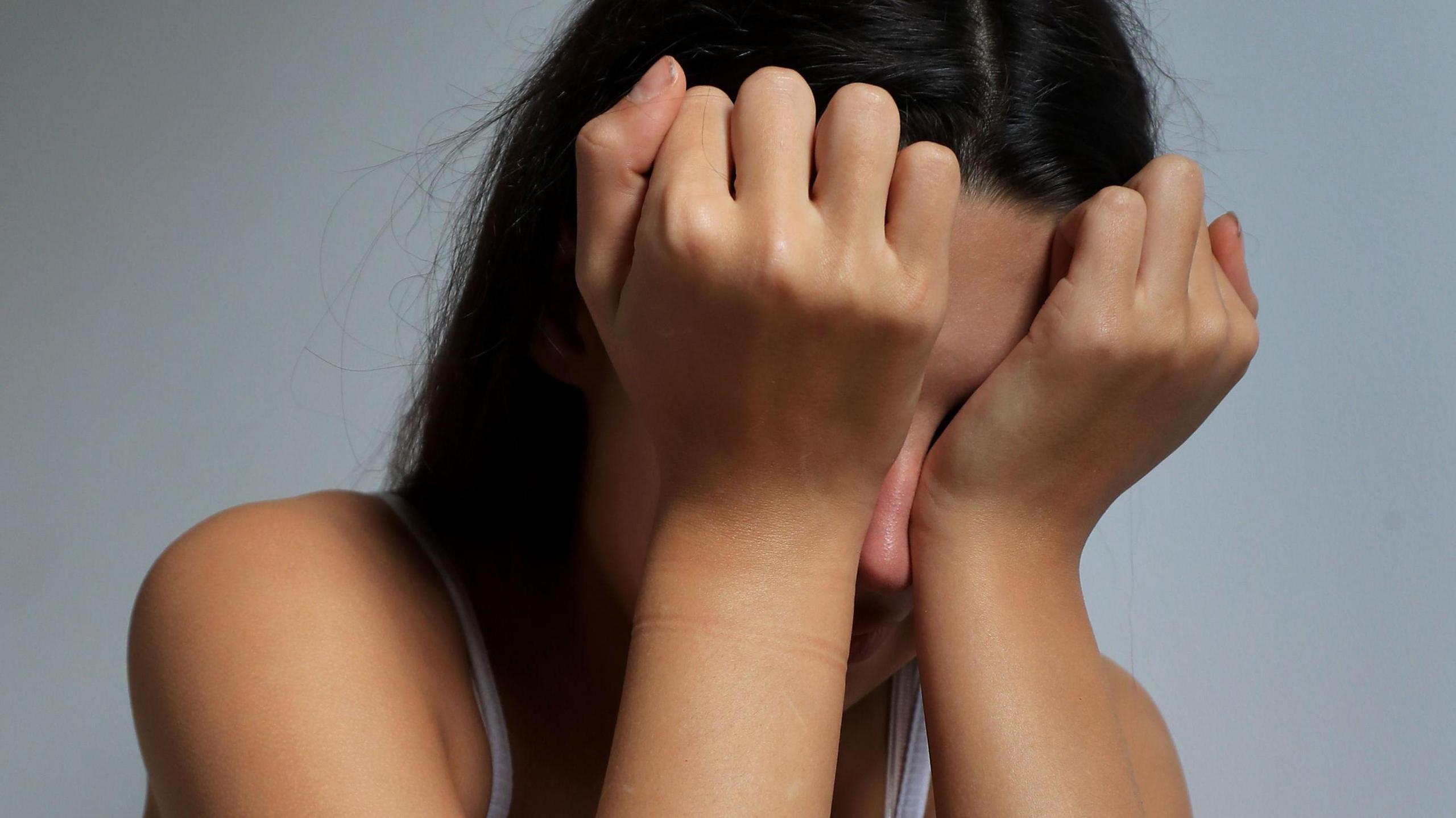 A teenage girl with olive skin and dark hair covers her eyes with her face in a crying pose.