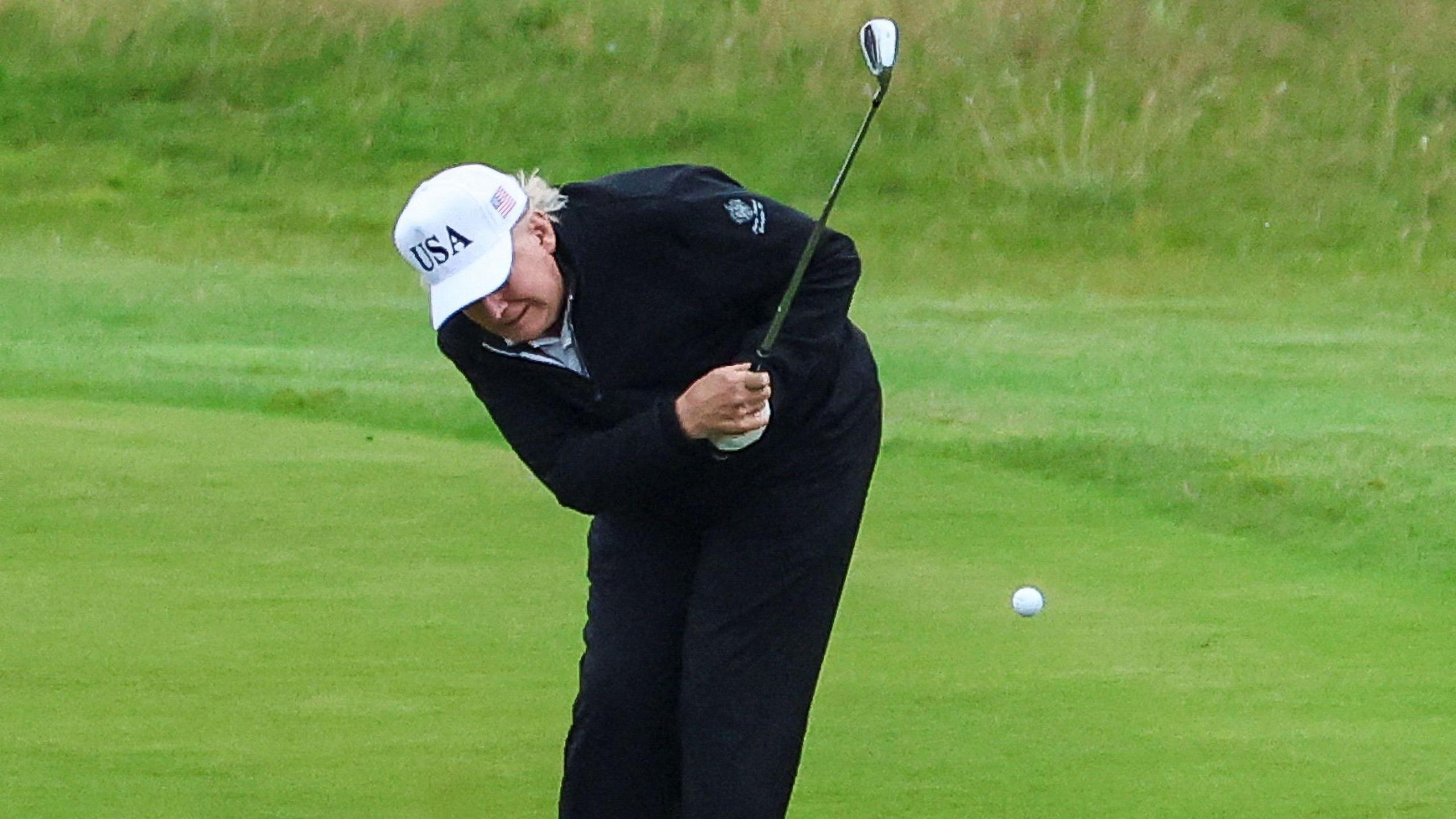 Donald Trump, wearing navy trousers and a navy jacket with a white "USA" cap, is doubled over as he strikes a golf ball.