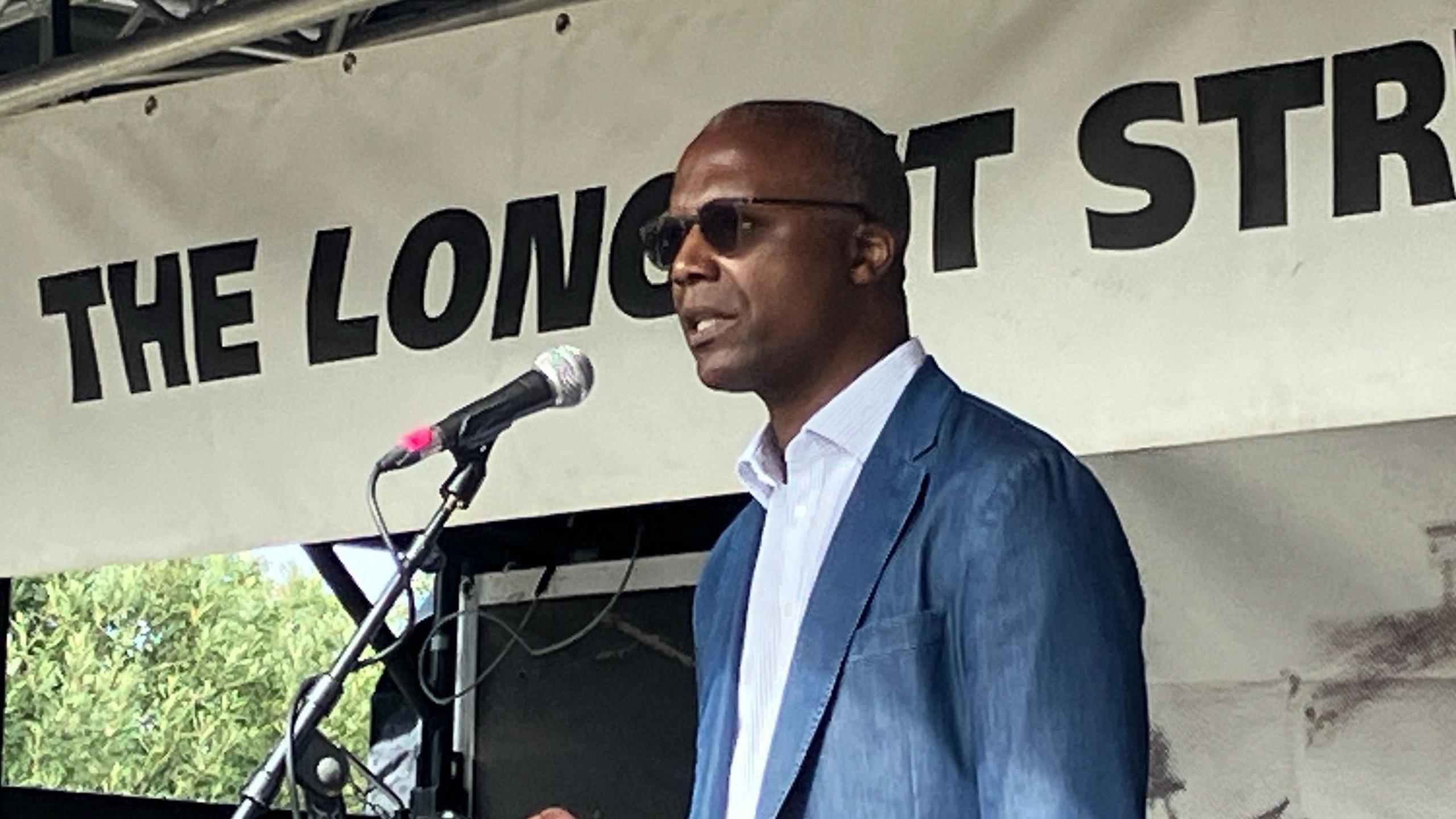 Patrick Roach speaks into a microphone on stage at the rally, he is reading from a mobile phone and wearing a white shirt, jacket and sunglasses