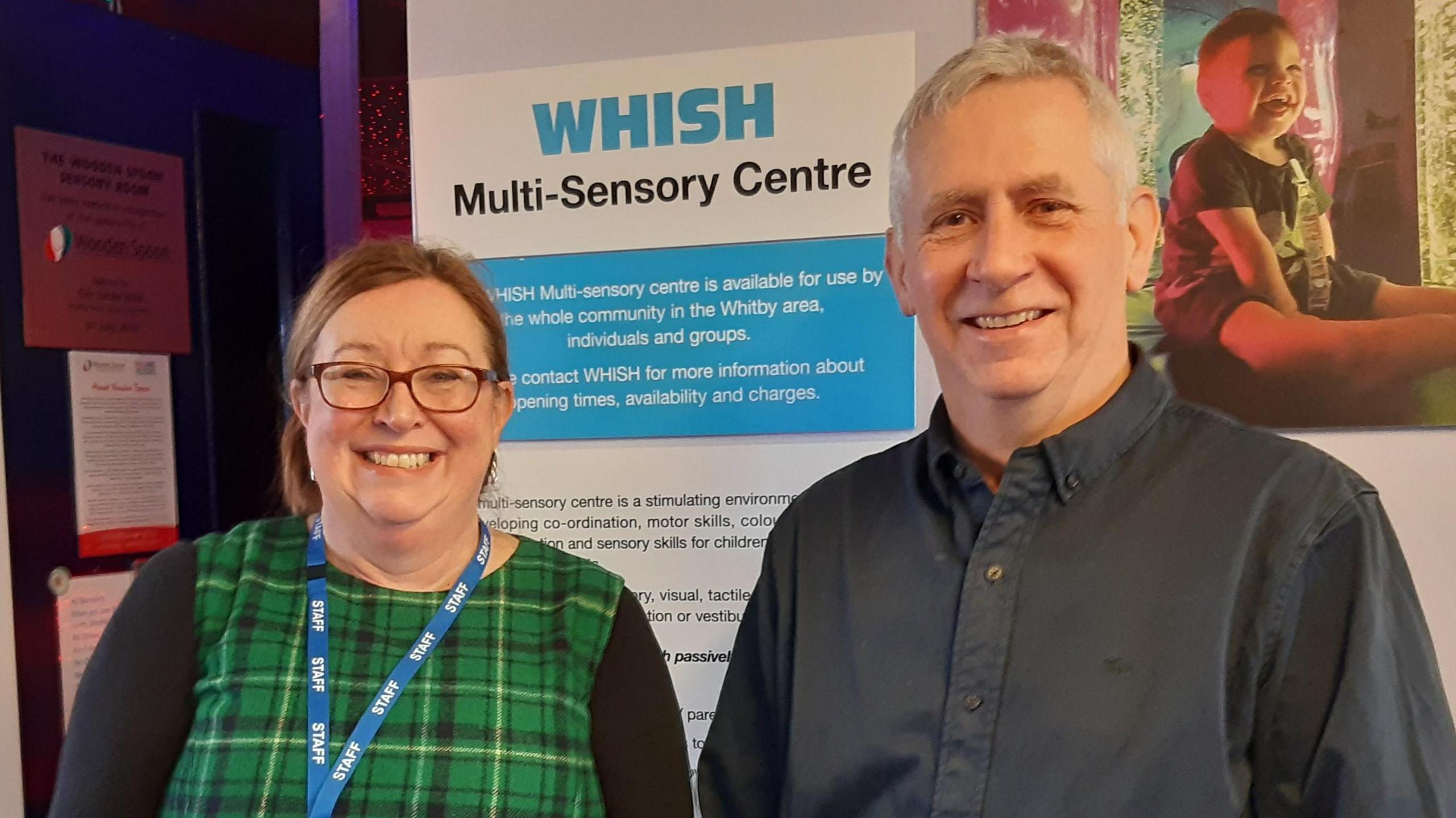 A woman wearing a green tartan dress and a blue staff lanyard stands smiling beside a man with grey hair wearing a dark shirt. Behind them is a picture of a giggling toddler in a multi-sensory room full of bright lights, beside a sign reading "WHISH multi-sensory centre".