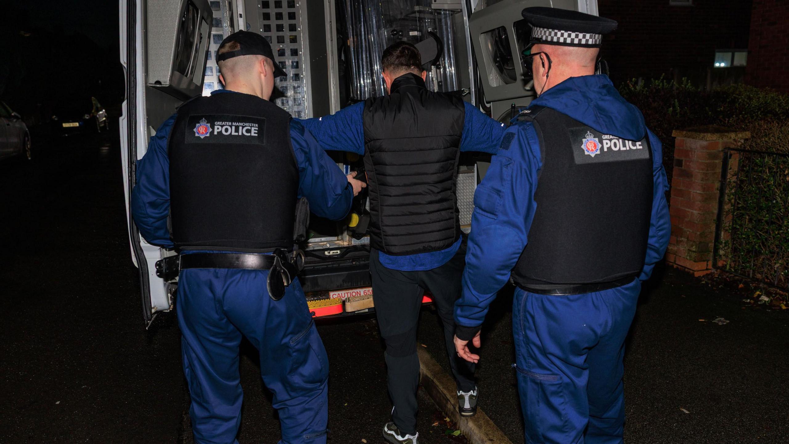 A man wearing  a blue long sleeved top under a black bodywarmer and black jogging bottoms is put in the back of a police van by two officers