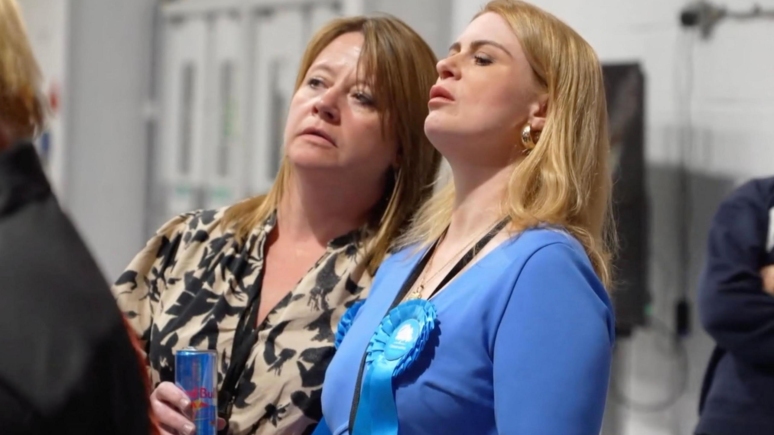 Two Conservative party members, one wearing a blue rosette look shocked as they watch votes being counted on election night in Norwich.