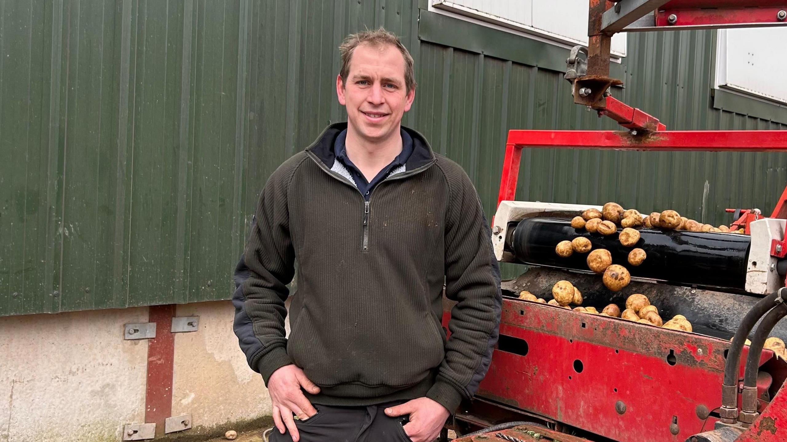 Alastair Heath wearing a green jumper stood next to a red machine which is sorting potatoes