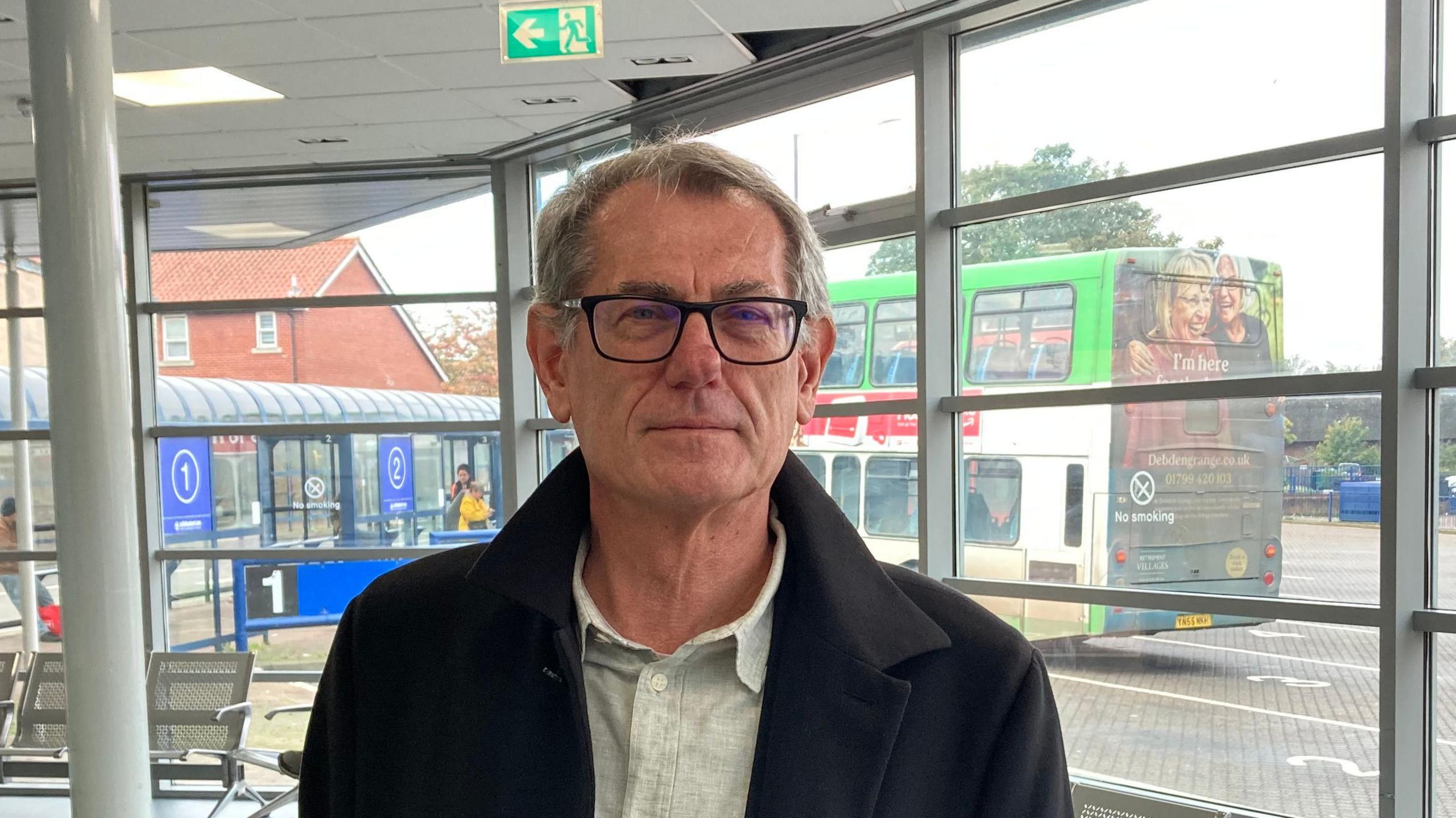 Cliff Waterman smiles at the camera while standing inside a bus station. He has short grey hair and is wearing black glasses with a black coat and off white shirt underneath. Behind him outside a parked double decker bus can be seen.