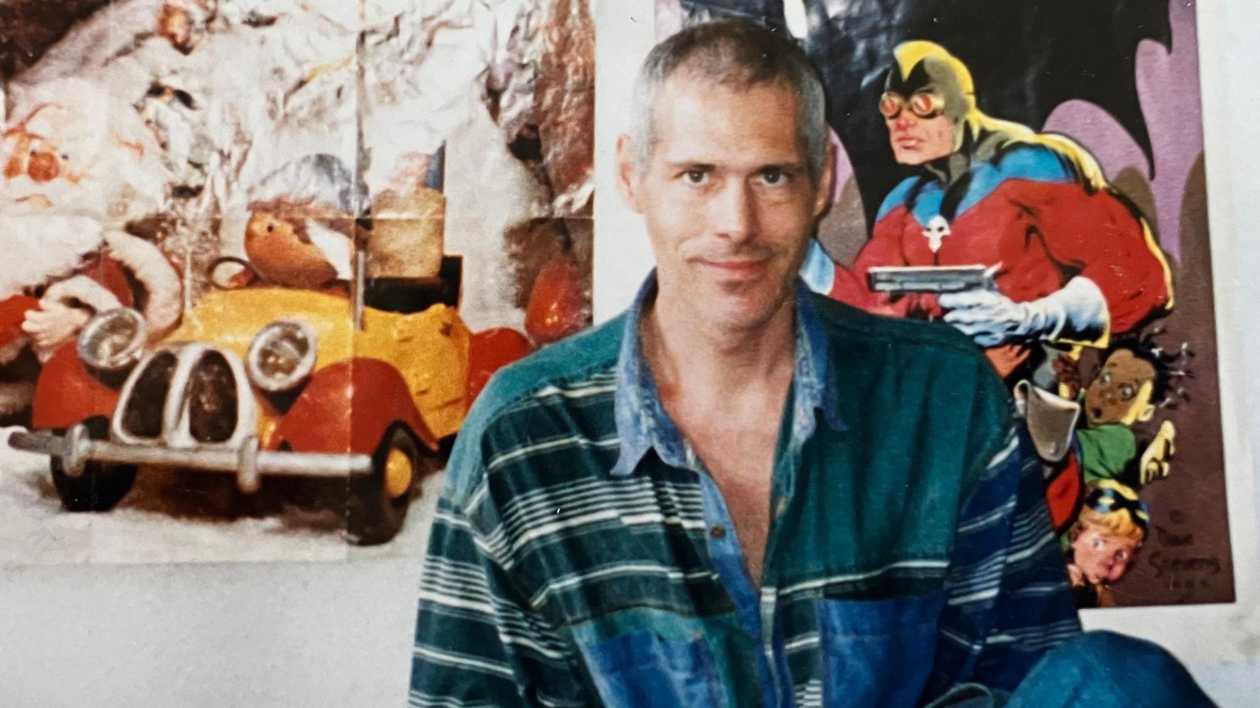 Nigel sitting in what appears to be a bedroom. He appears to be in his late 30s or early 40s. He is wearing an oversized green and blue shirt. There are children's television programme posters on the wall behind him.